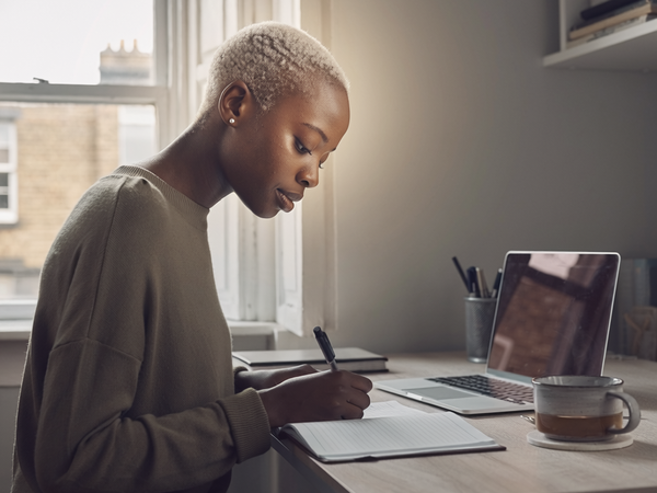 Black woman at home, focused on a notebook in natural daylight.