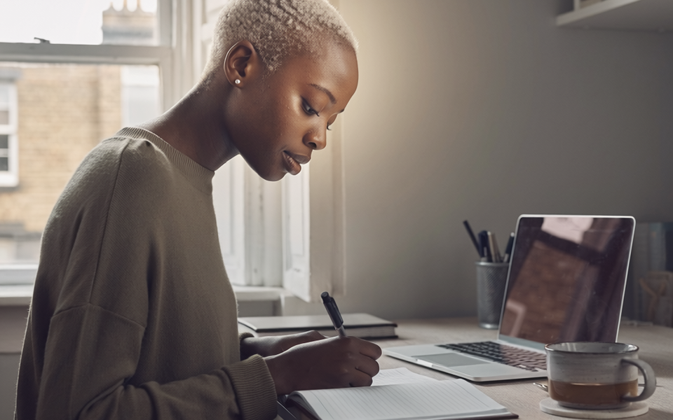 Black woman at home, focused on a notebook in natural daylight.