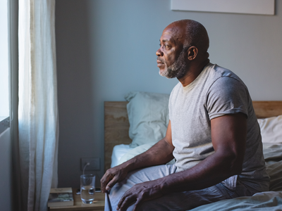 Black man in his fifties sitting on the edge of a bed in a calm London bedroom in the morning.