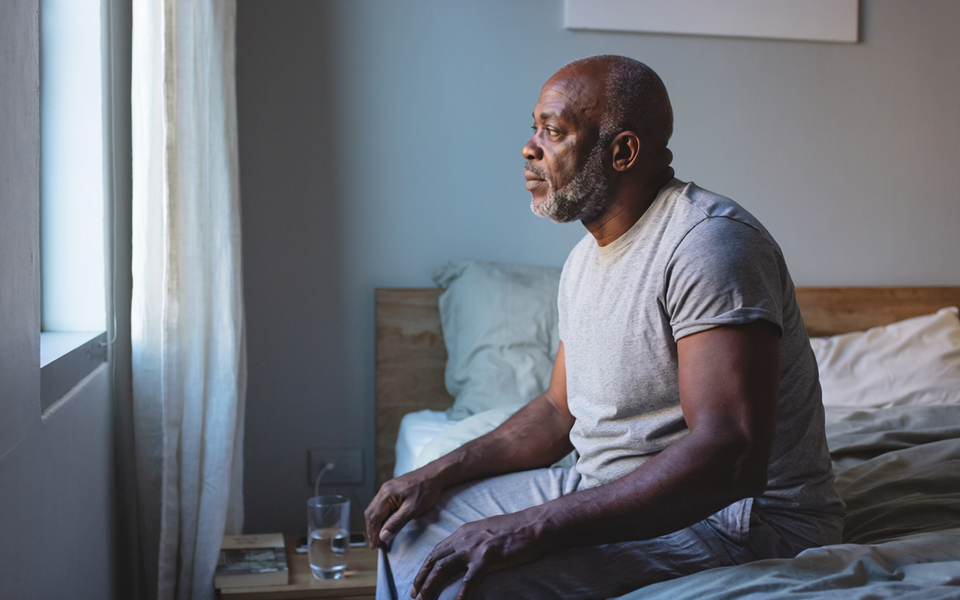 Black man in his fifties sitting on the edge of a bed in a calm London bedroom in the morning.