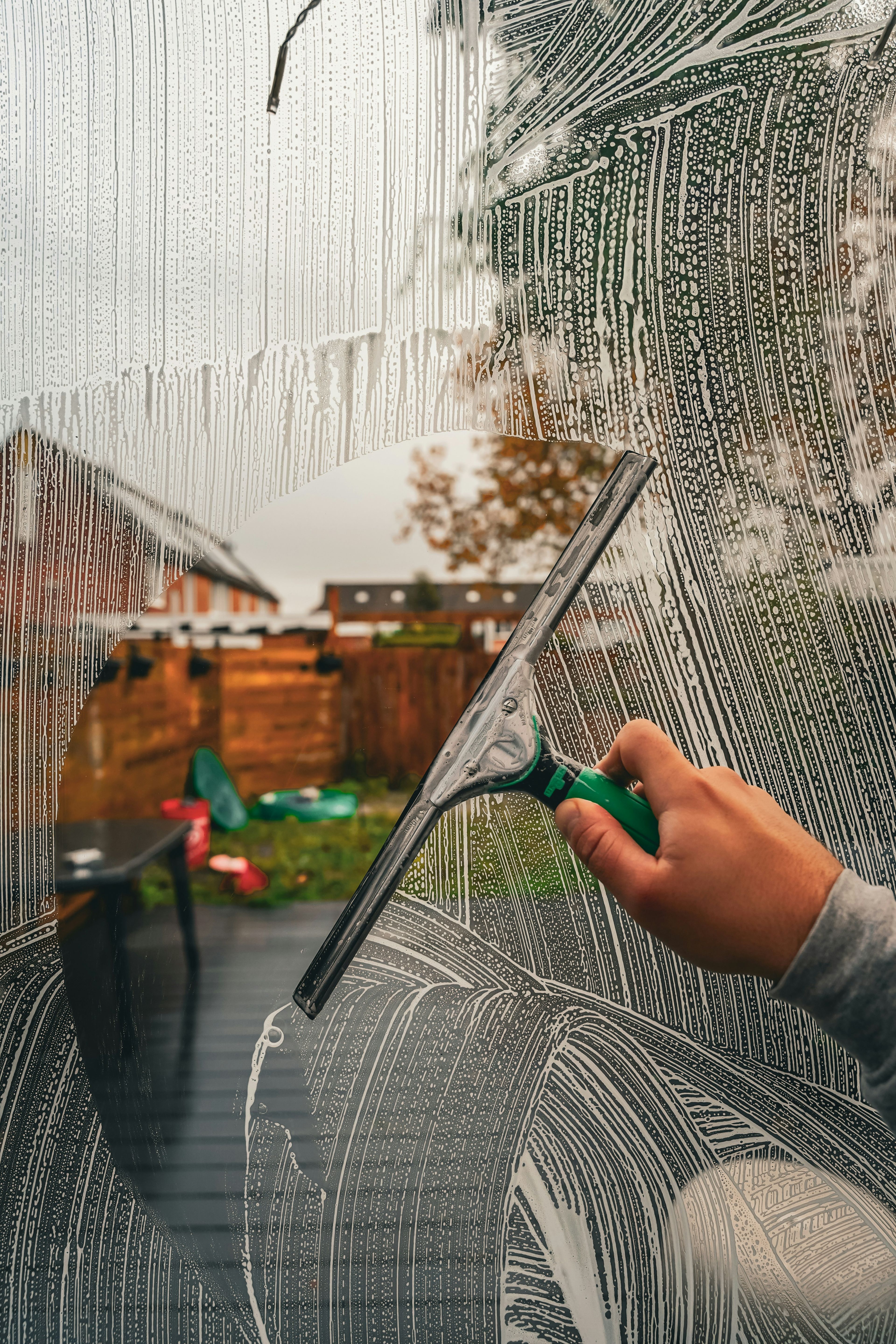 A hand using a squeegee to clean a soapy window, revealing a backyard with blurred houses, trees, and garden furniture on a cloudy day.
