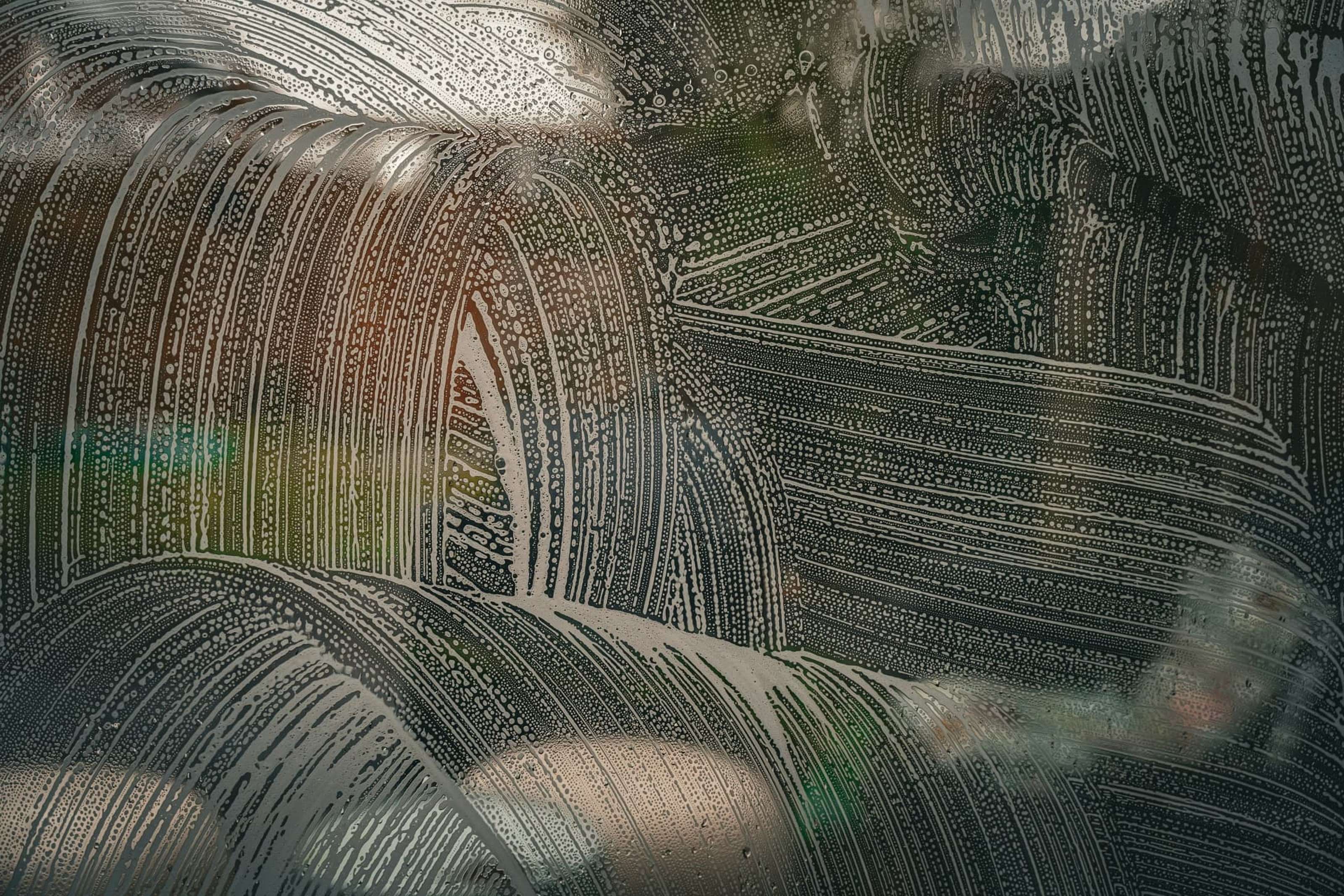 Streaks and droplets of water on a glass surface create an intricate pattern, with a blurred green and brown background visible through the glass.