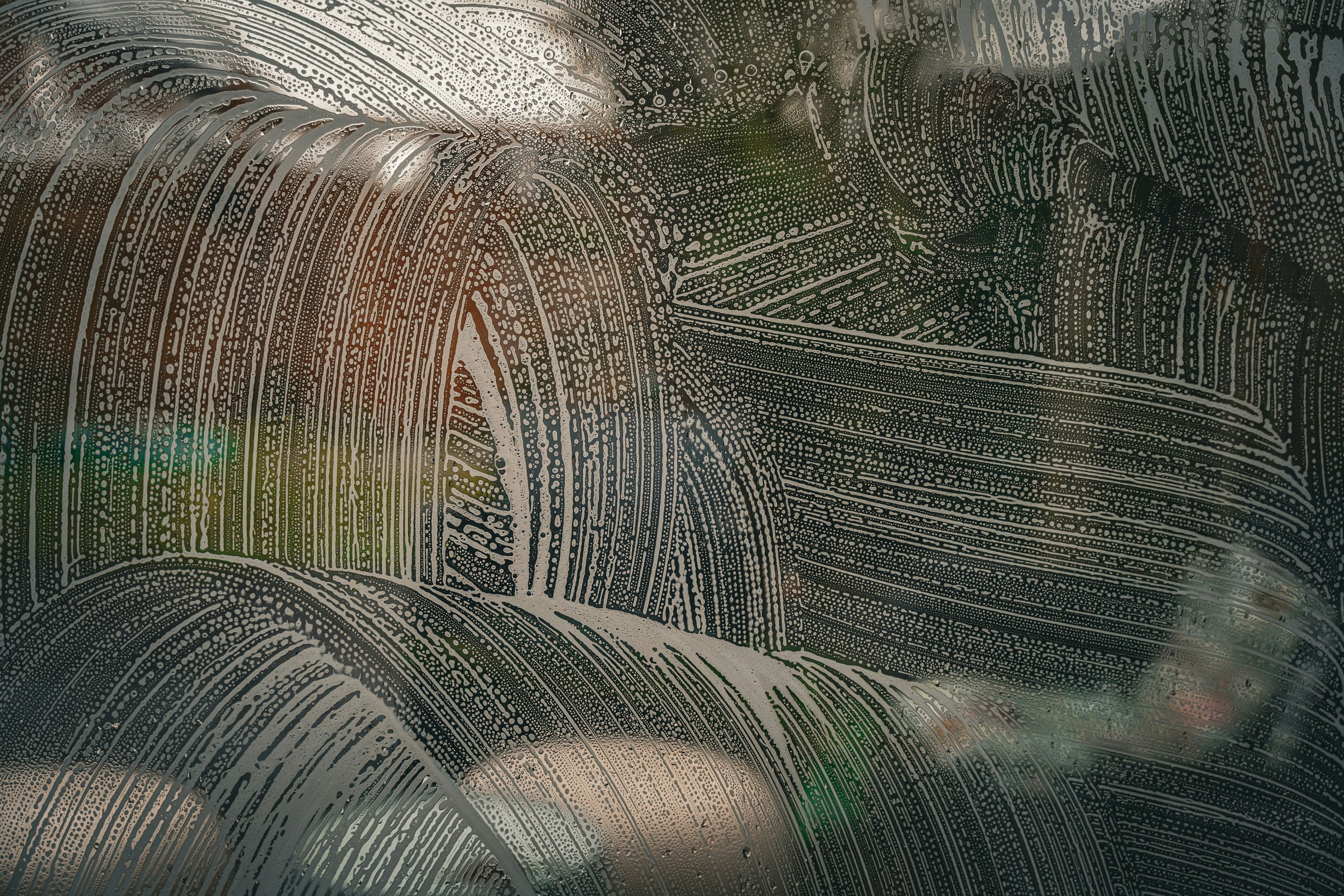 Streaks and droplets of water on a glass surface create an intricate pattern, with a blurred green and brown background visible through the glass.