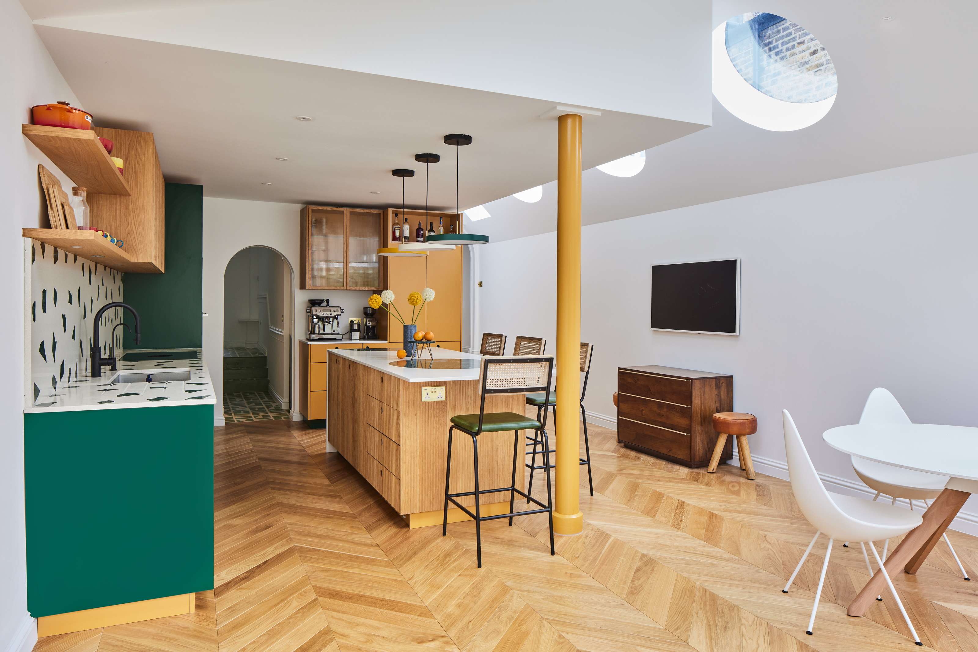 Bright open-plan kitchen and dining area with green cabinetry, wooden island and stools, yellow column, skylights, and herringbone wood floor.