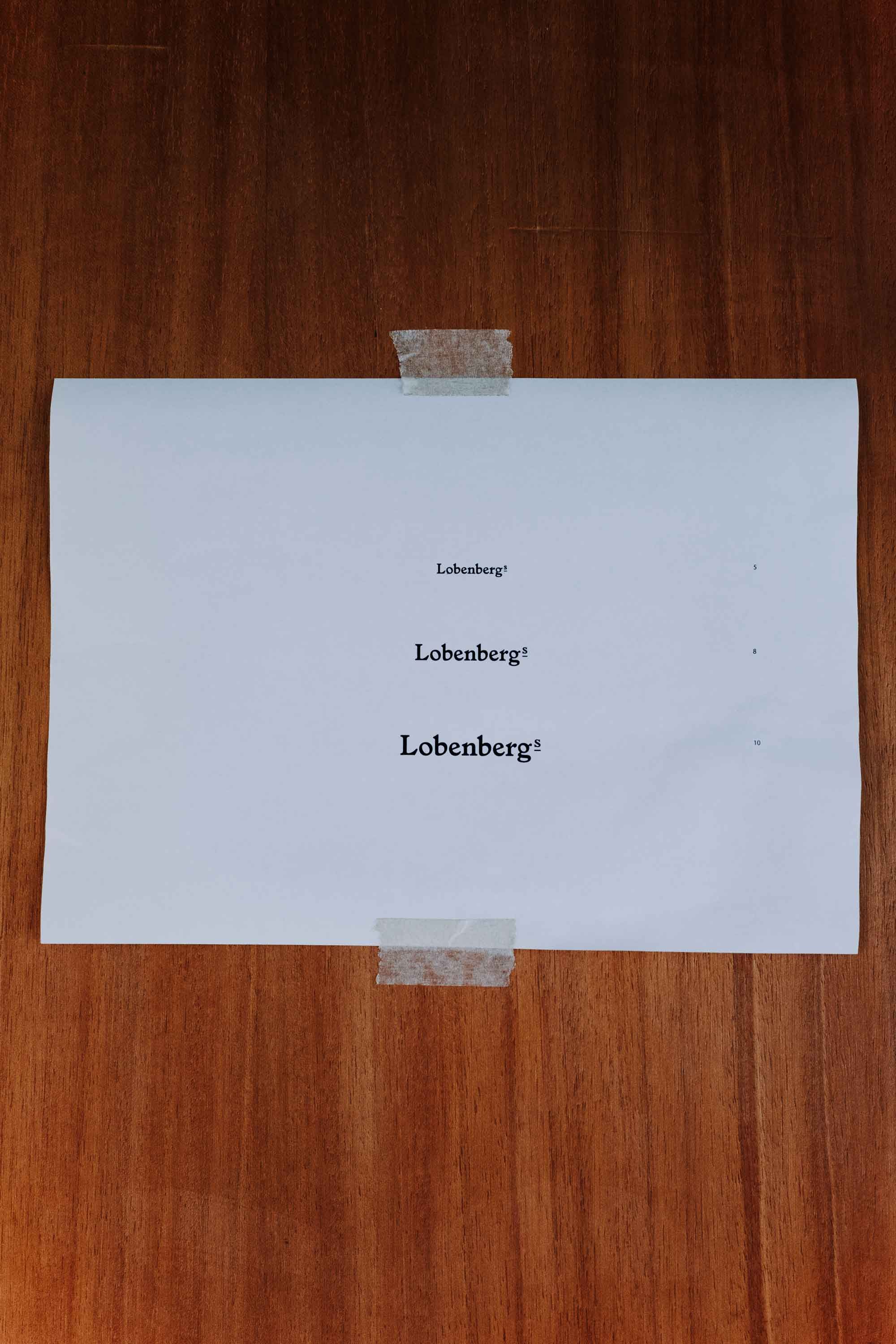 Testausdrucke für eine Wortmarke »Lobenbergs« auf einem Zettel an einer Holzfurnierwand
