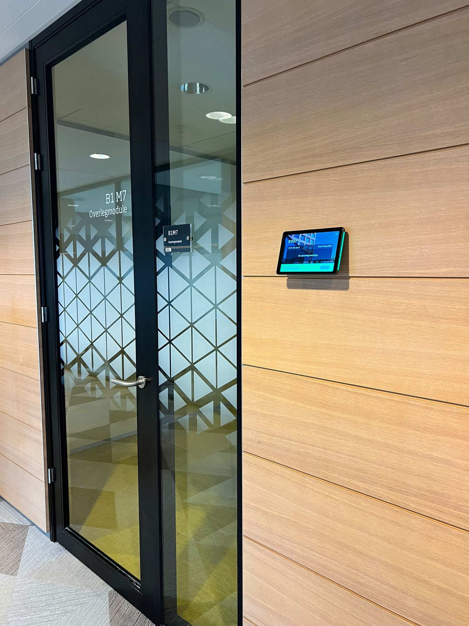 A modern office door with a glass panel featuring a geometric pattern and a digital display mounted on the wood-paneled wall beside it.