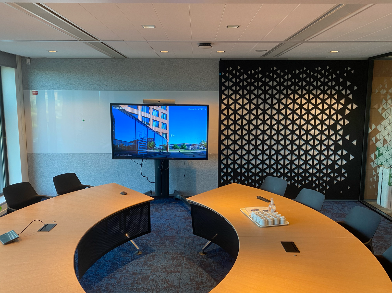 Modern conference room with curved wooden tables, black chairs, a large screen displaying a building, and patterned wall decor.