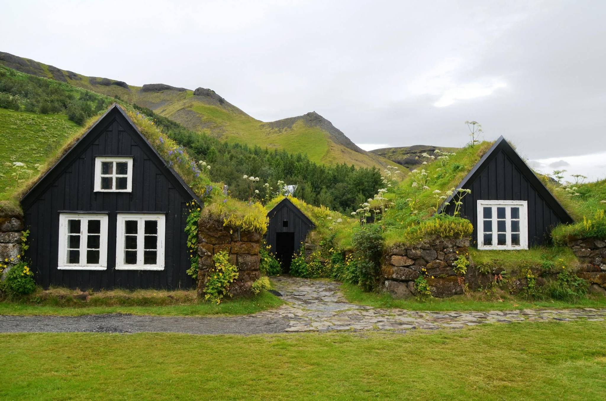 Two black wooden houses with grass-covered roofs, white windows, and stone paths, set against a backdrop of green hills and overcast sky.