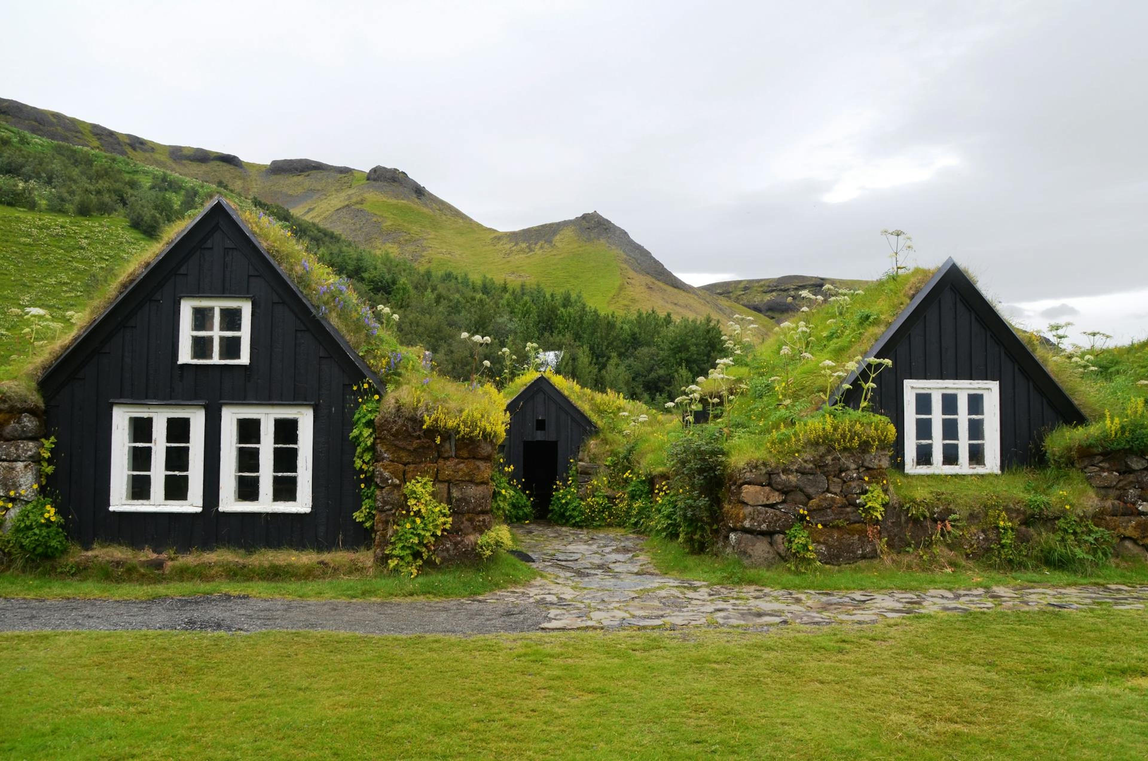 Two black wooden houses with grass-covered roofs, white windows, and stone paths, set against a backdrop of green hills and overcast sky.