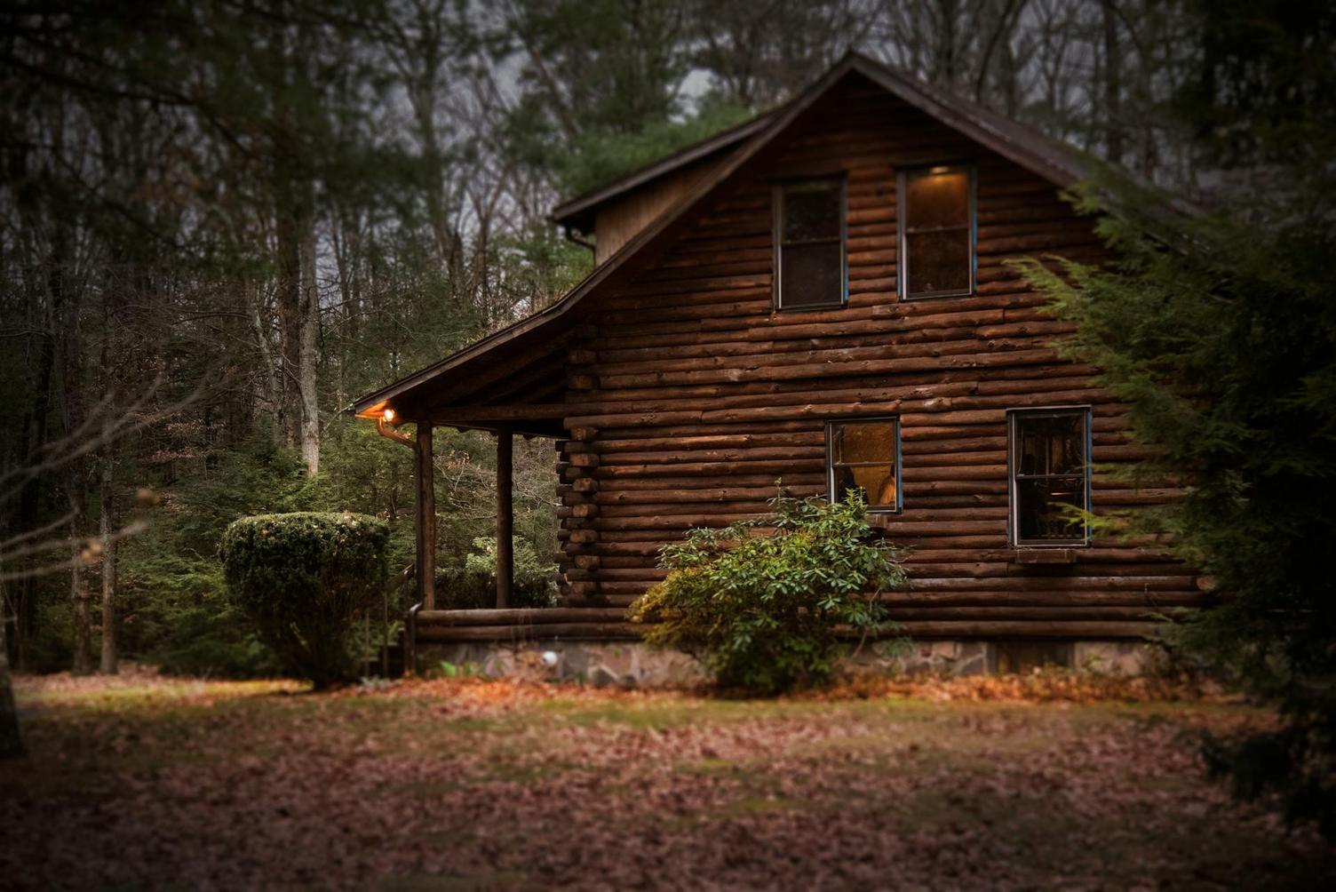 A rustic log cabin surrounded by trees, with a small porch and warm lights glowing from the windows, set in a tranquil forest setting.