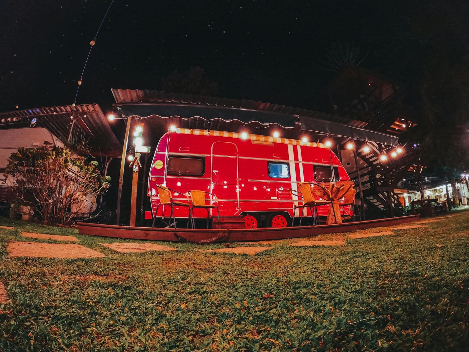 A red camper van is parked under string lights at night, surrounded by grass and a wooden deck with chairs.