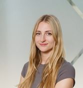 Woman with long blonde hair in a gray shirt, arms crossed, smiling slightly against a neutral background.
