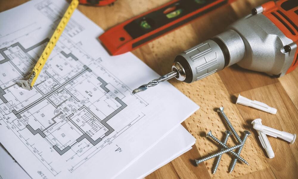 Blueprints on a table with a drill, screws, wall plugs, a tape measure, and a level, suggesting a construction or renovation project.