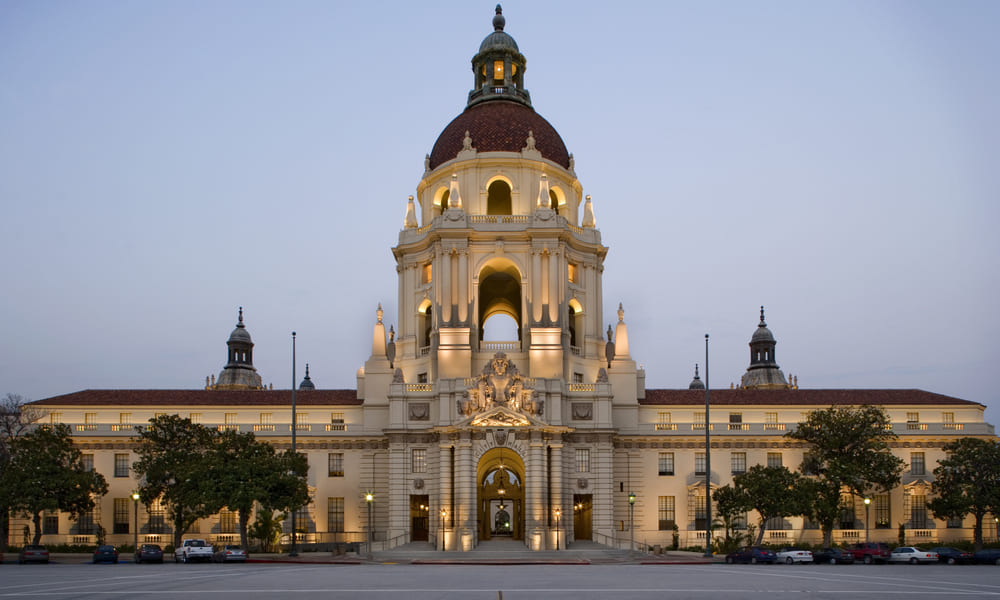 Illuminated building with a large dome and arched entrance, flanked by two smaller domes, set against a twilight sky.