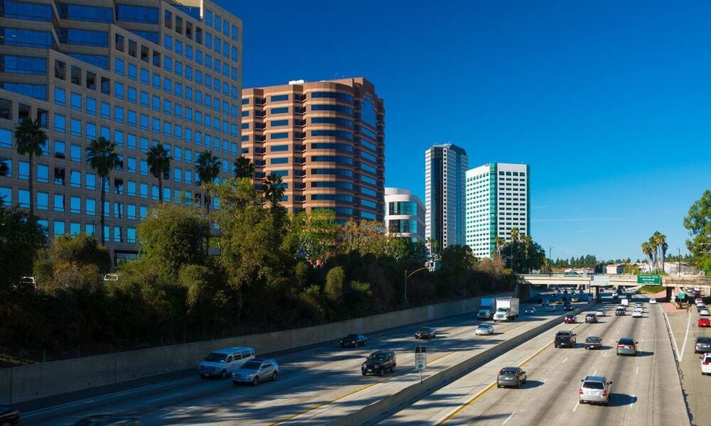 Busy highway with cars and trucks, lined by tall modern buildings and greenery under a clear blue sky.