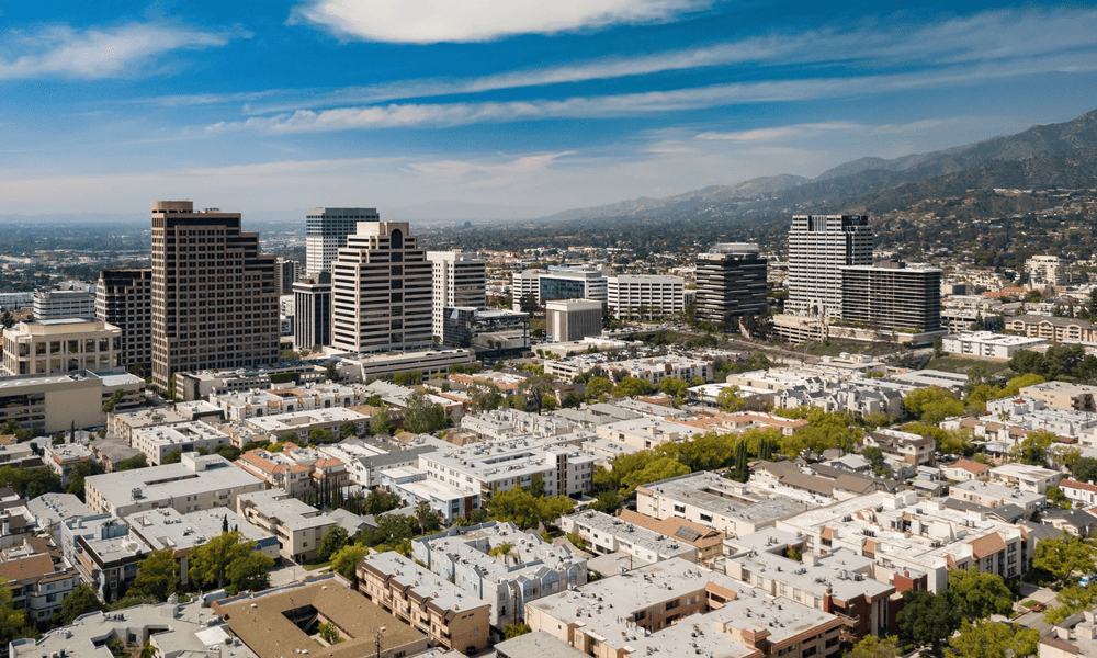 Aerial view of a cityscape with modern skyscrapers, residential buildings, and mountains under a blue sky with scattered clouds.