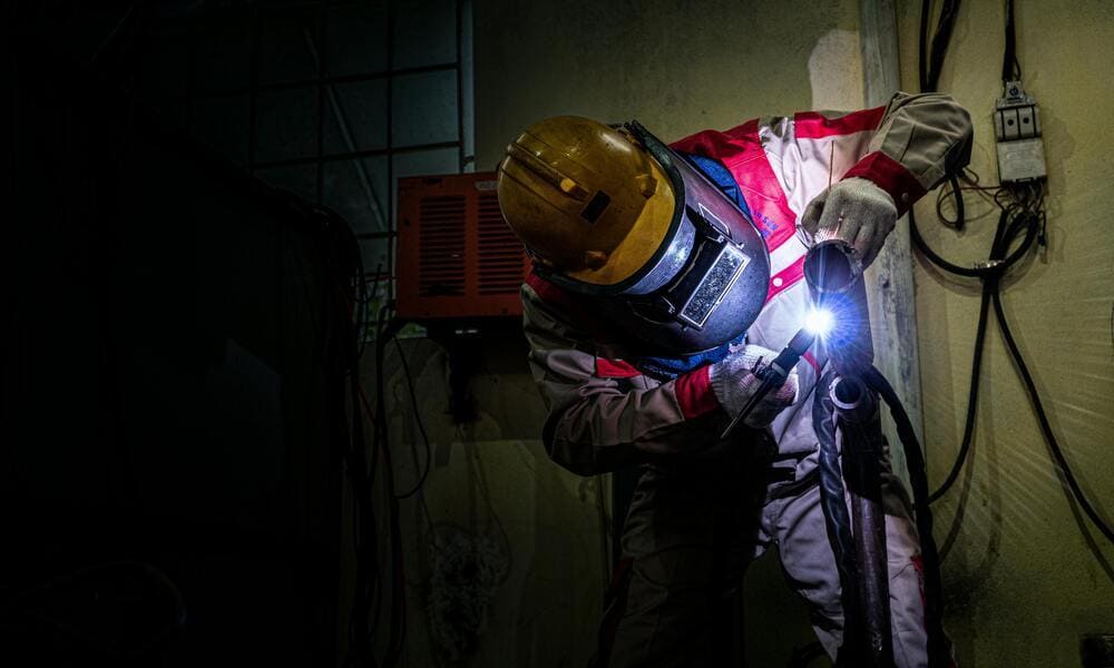 A welder in protective gear works with a bright torch in a dimly lit industrial setting.