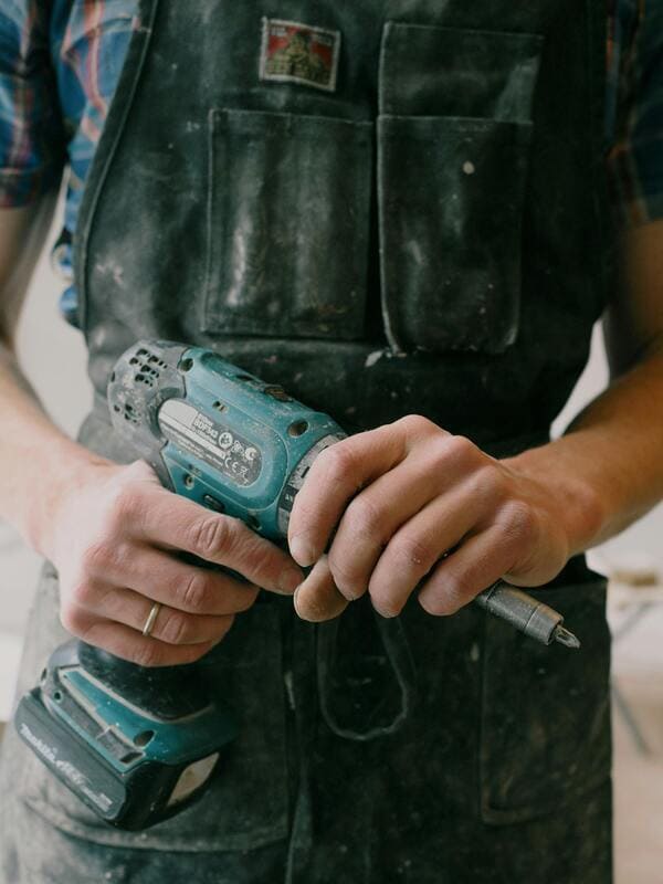 Person in a dusty apron holding a cordless drill, wearing a plaid shirt underneath. Hands are slightly dusty, suggesting recent work.