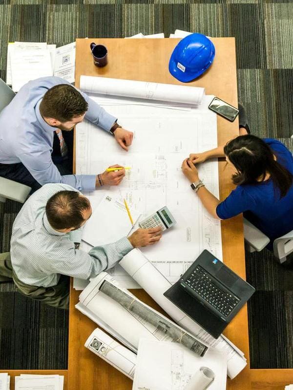 Three people collaborate over architectural plans at a table with a laptop, calculator, blueprints, and a blue hard hat.