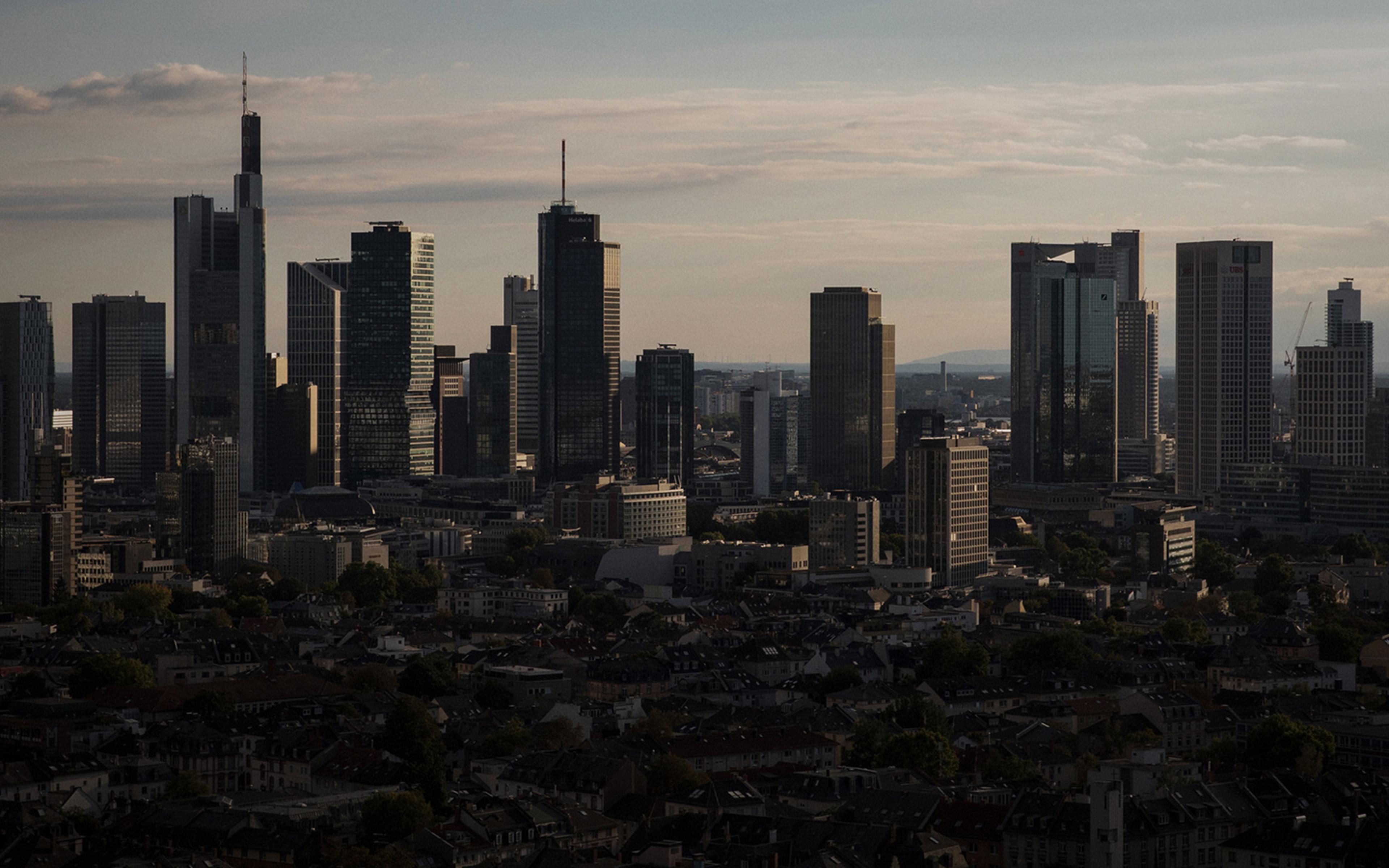 A city skyline with tall, modern skyscrapers under a cloudy sky, casting shadows over the urban landscape below.