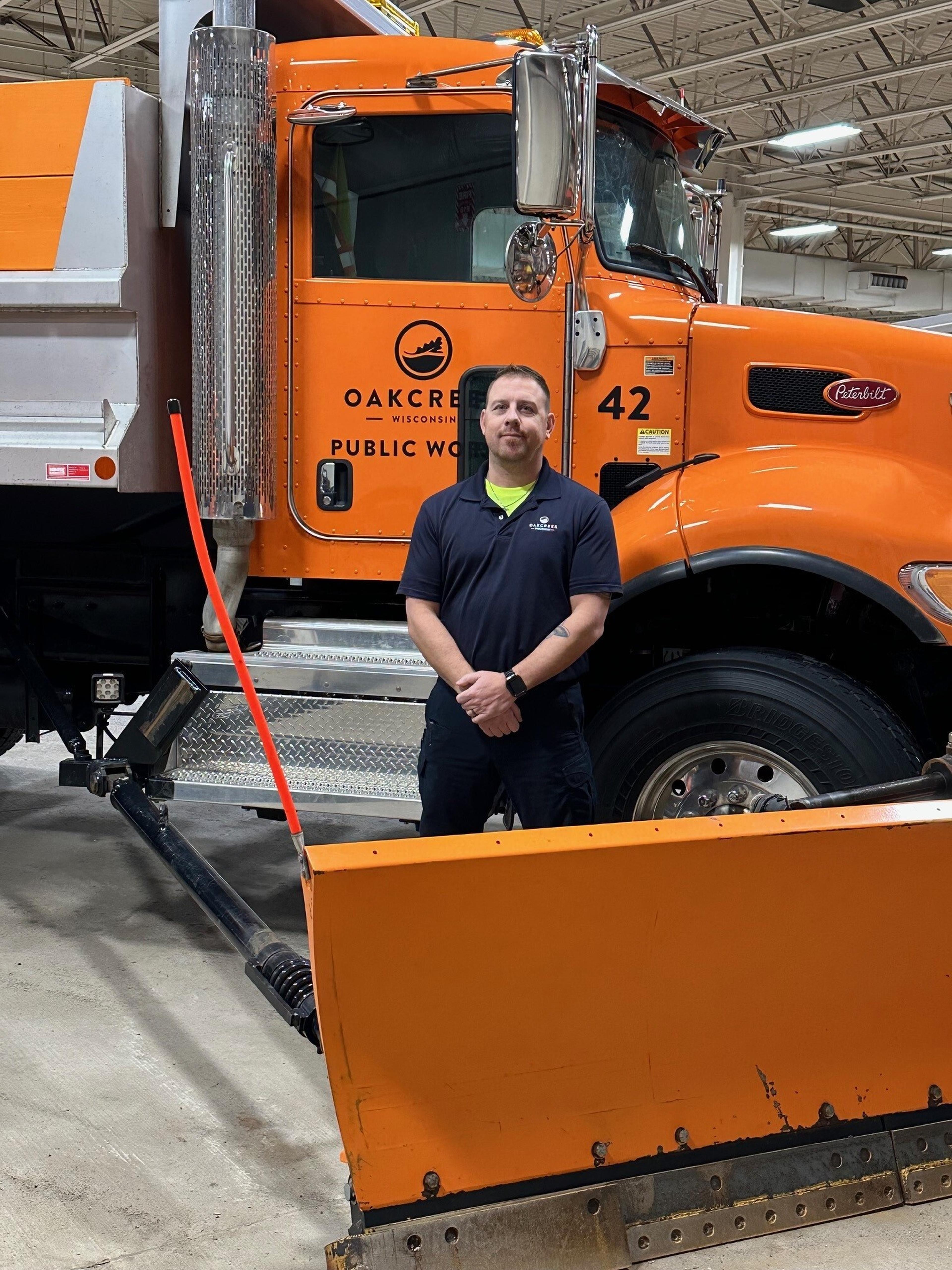 Man standing in front of an orange Oak Creek public works truck with a snowplow inside a garage.