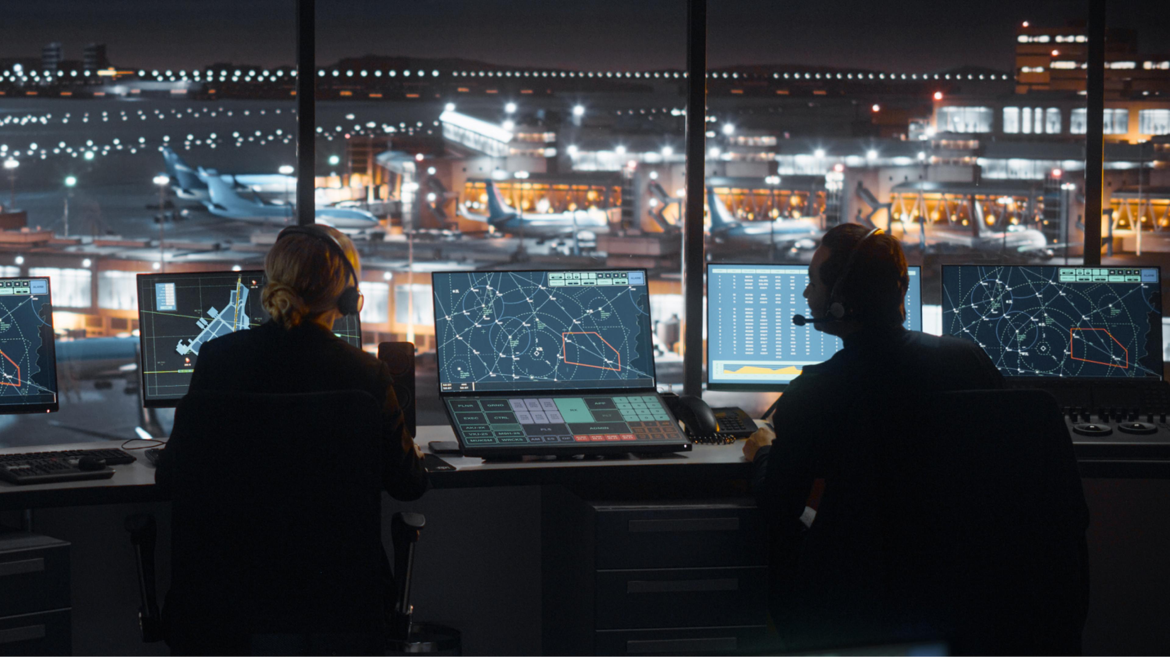 Two air traffic controllers wearing headsets monitor flight paths on multiple screens in a control tower overlooking an airport at night.
