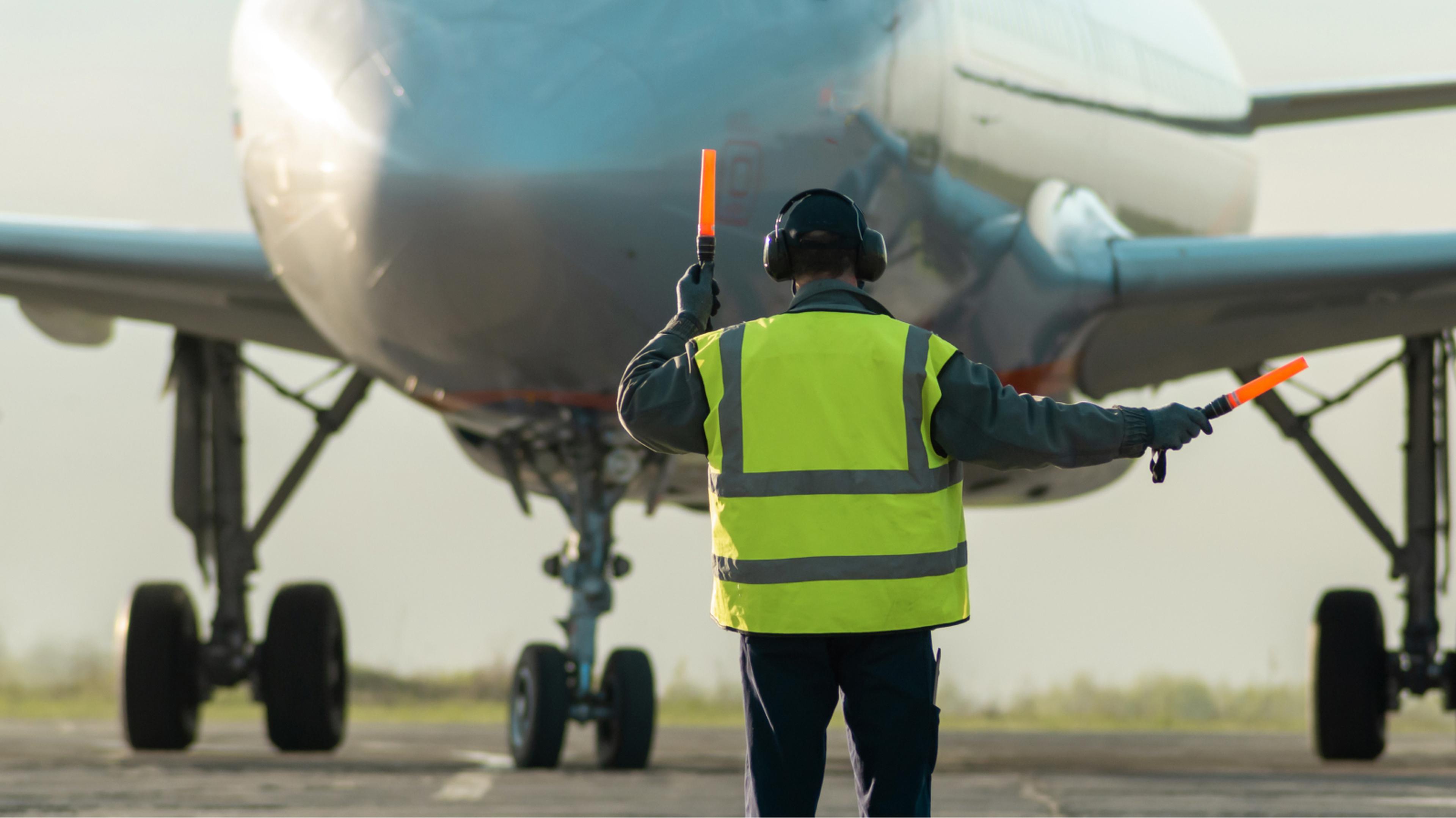 Airport ground crew member in a high-visibility vest uses orange wands to guide an approaching airplane on the runway.
