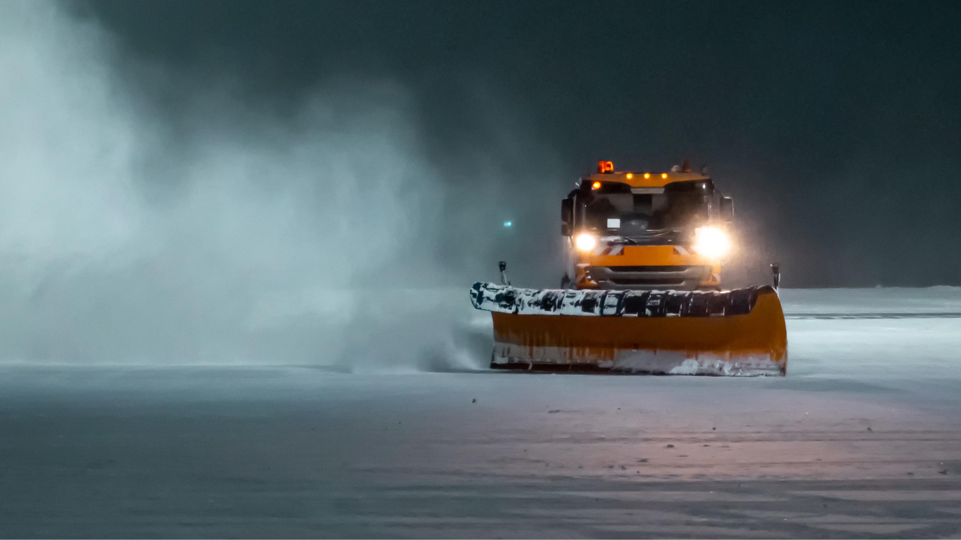 Snowplow clearing a snow-covered runway at night, with bright headlights illuminating the falling snow and creating a misty atmosphere.