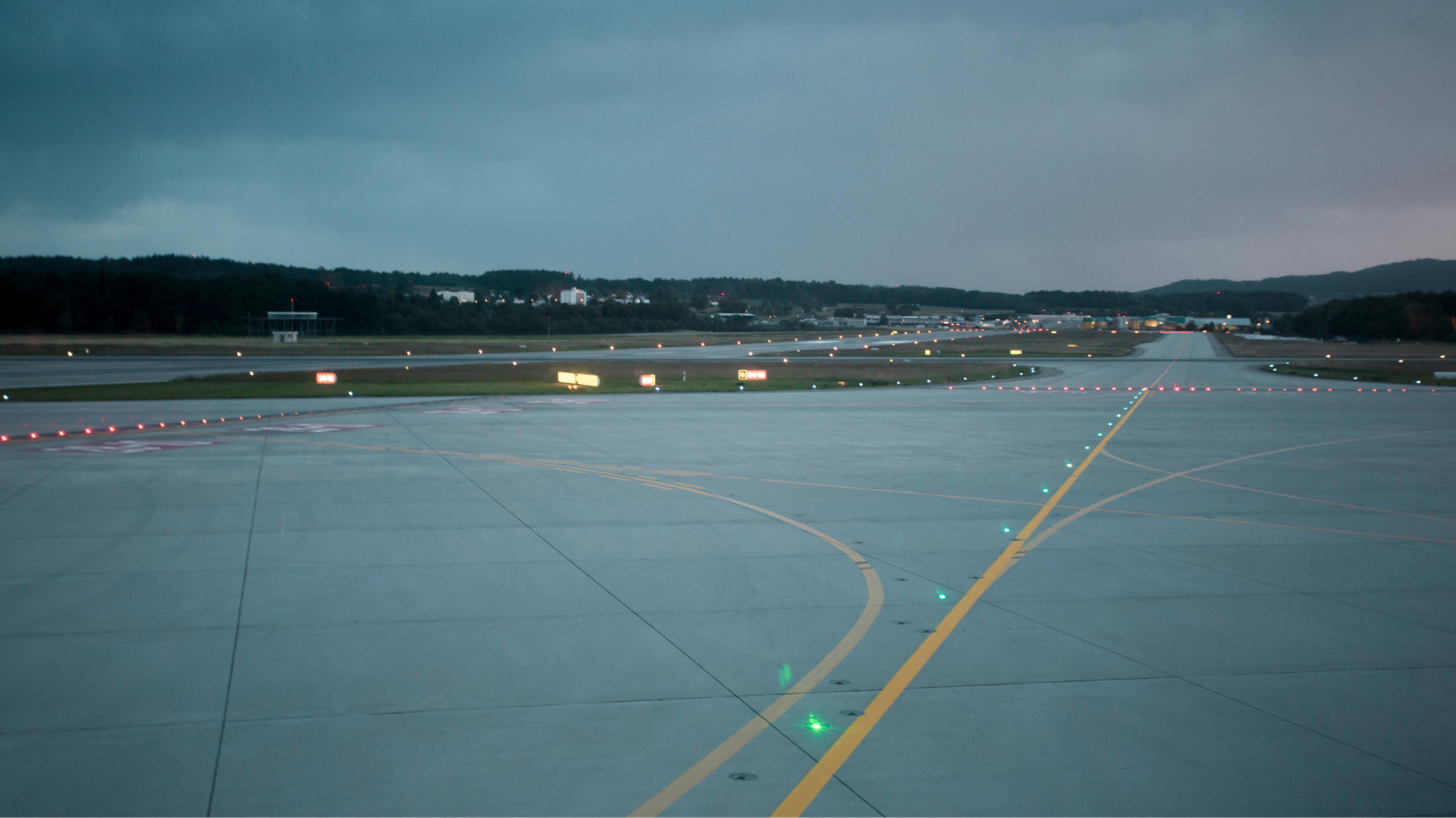 Airport runway at dusk with illuminated guiding lights and overcast sky; forested landscape in the background.