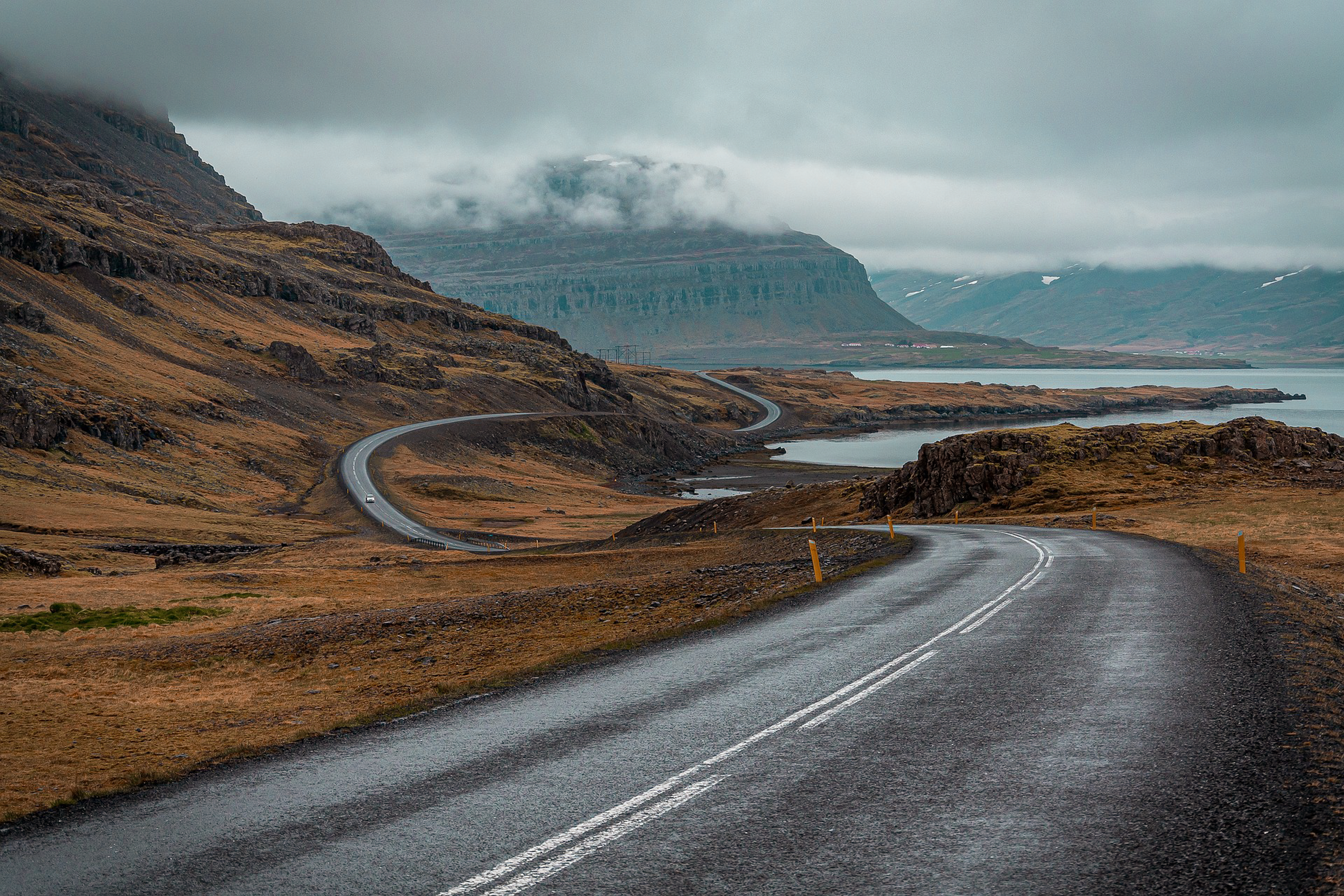 A winding road in a mountainous landscape under a cloudy sky, with distant cliffs and calm water alongside.