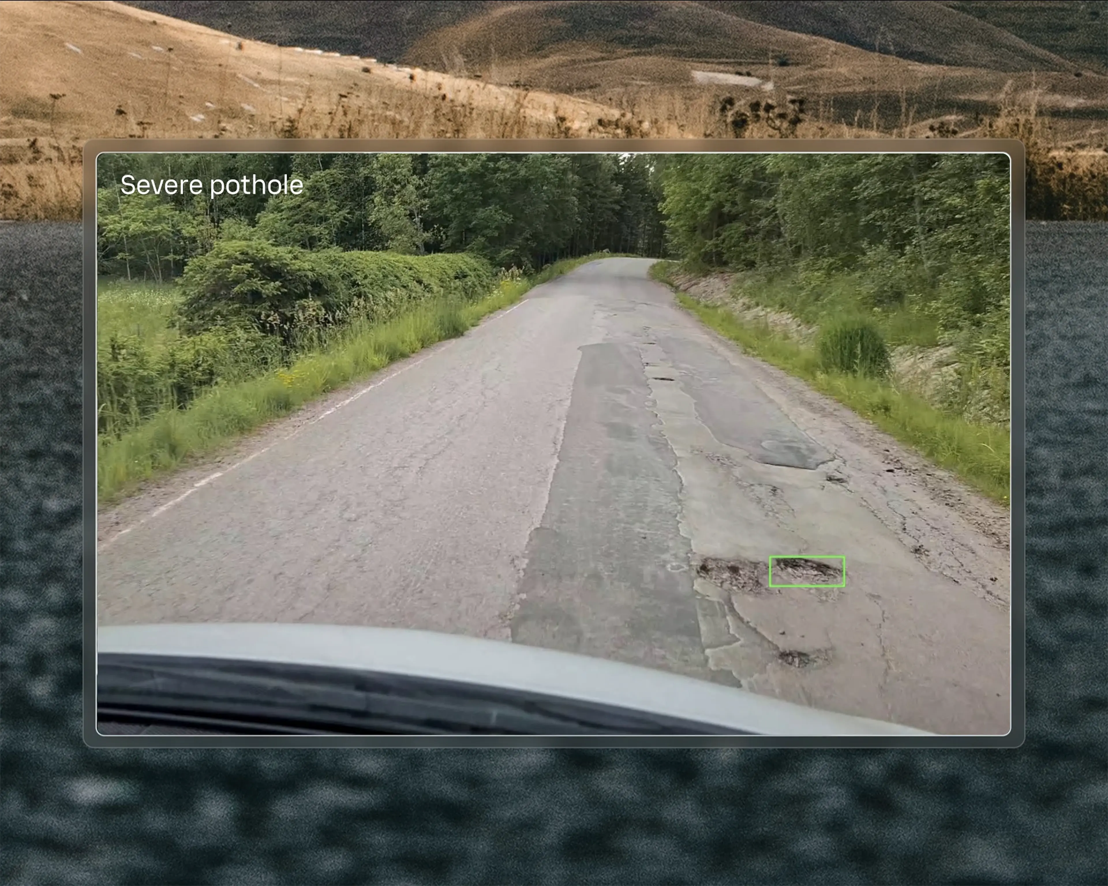 A rural road with a large, severe pothole in the center, surrounded by greenery and trees.