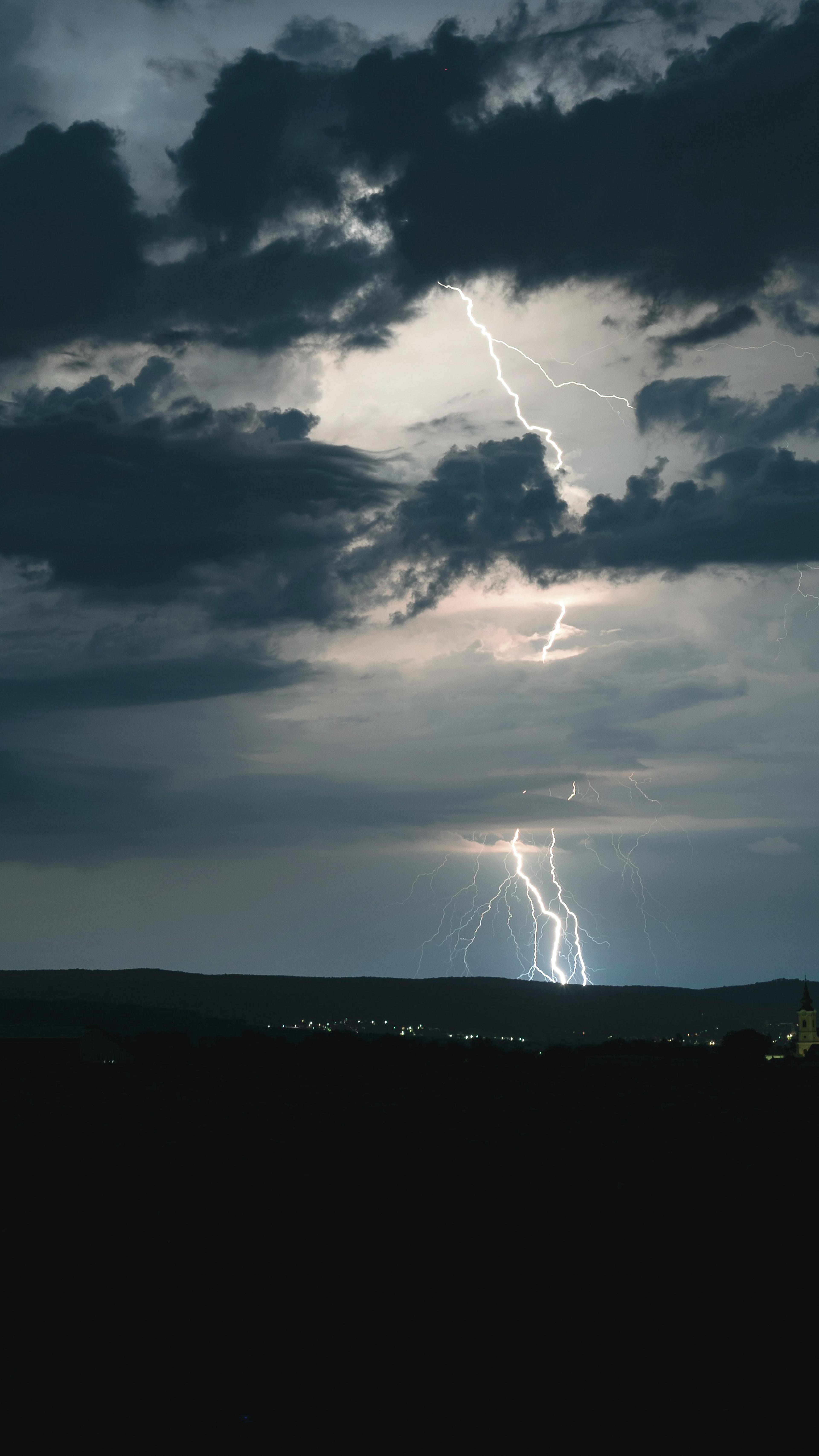 A vivid lightning bolt illuminates the dark, cloudy night sky above a distant cityscape, highlighting dramatic clouds.