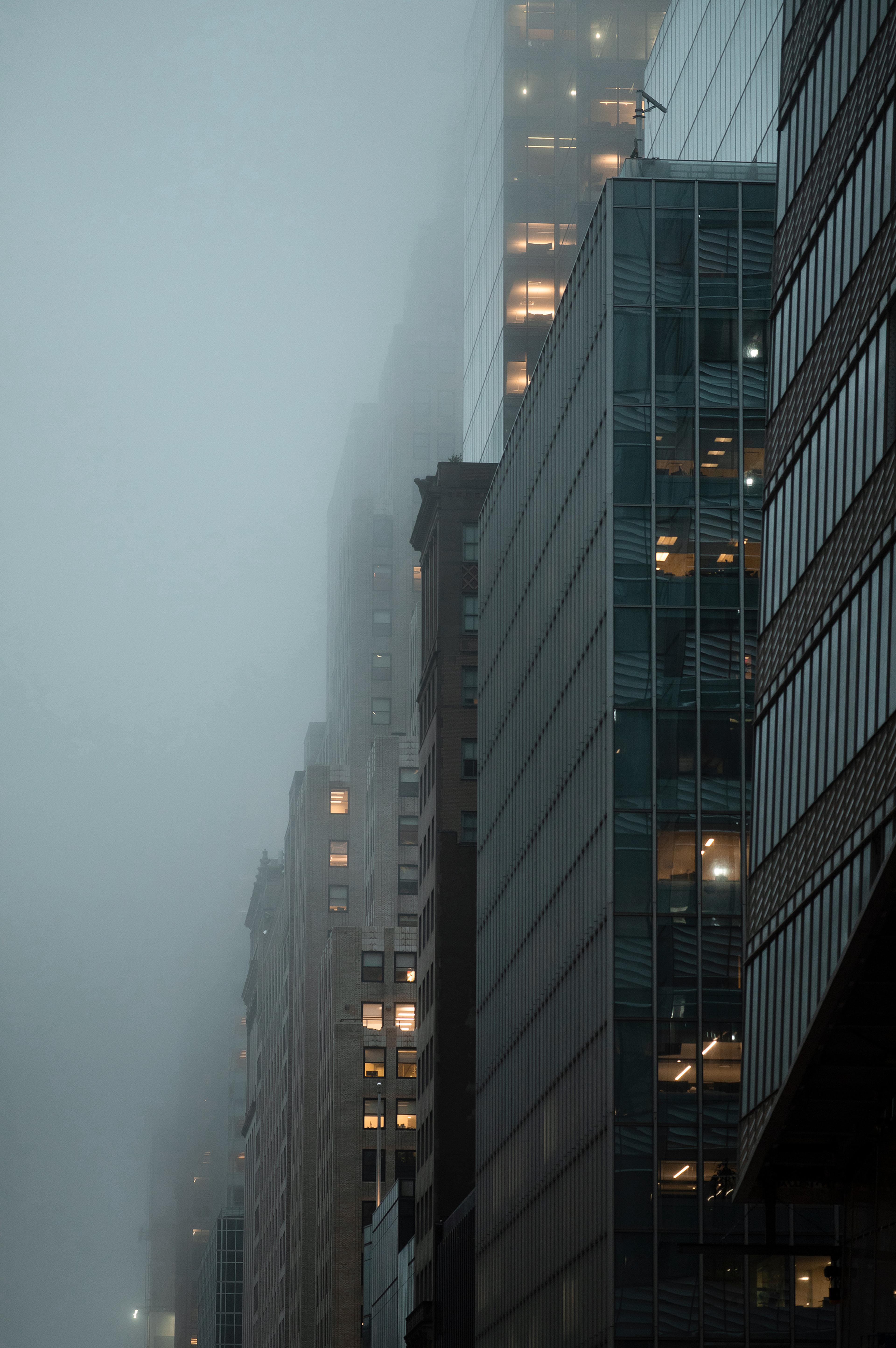 Foggy cityscape with tall buildings partially obscured, lit windows glowing softly. Modern architecture contrasts against the misty background.