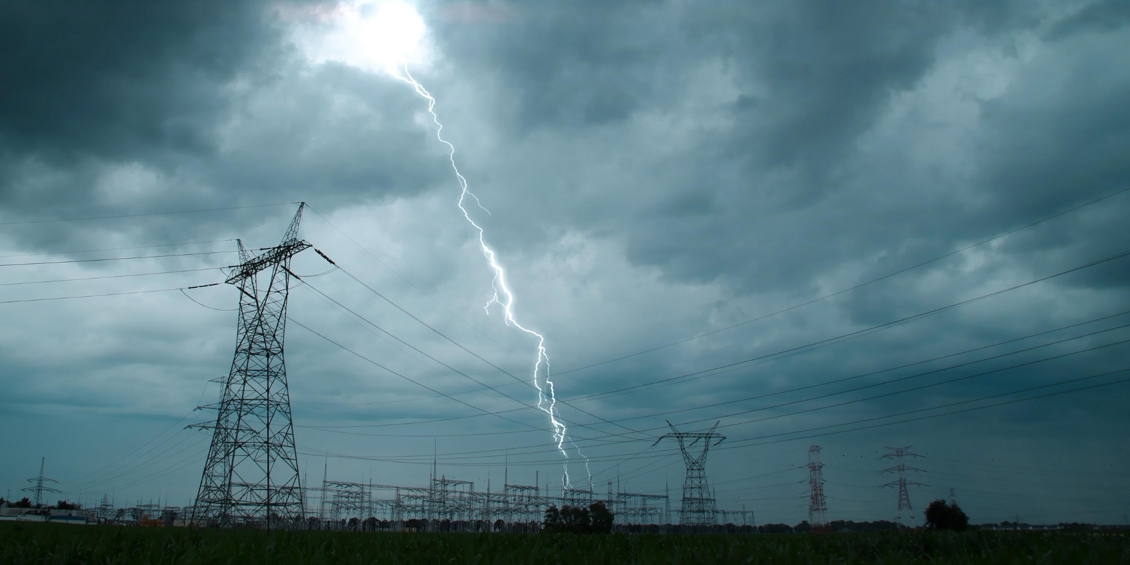 Lightning strikes near power lines under a dark, cloudy sky in an open field.