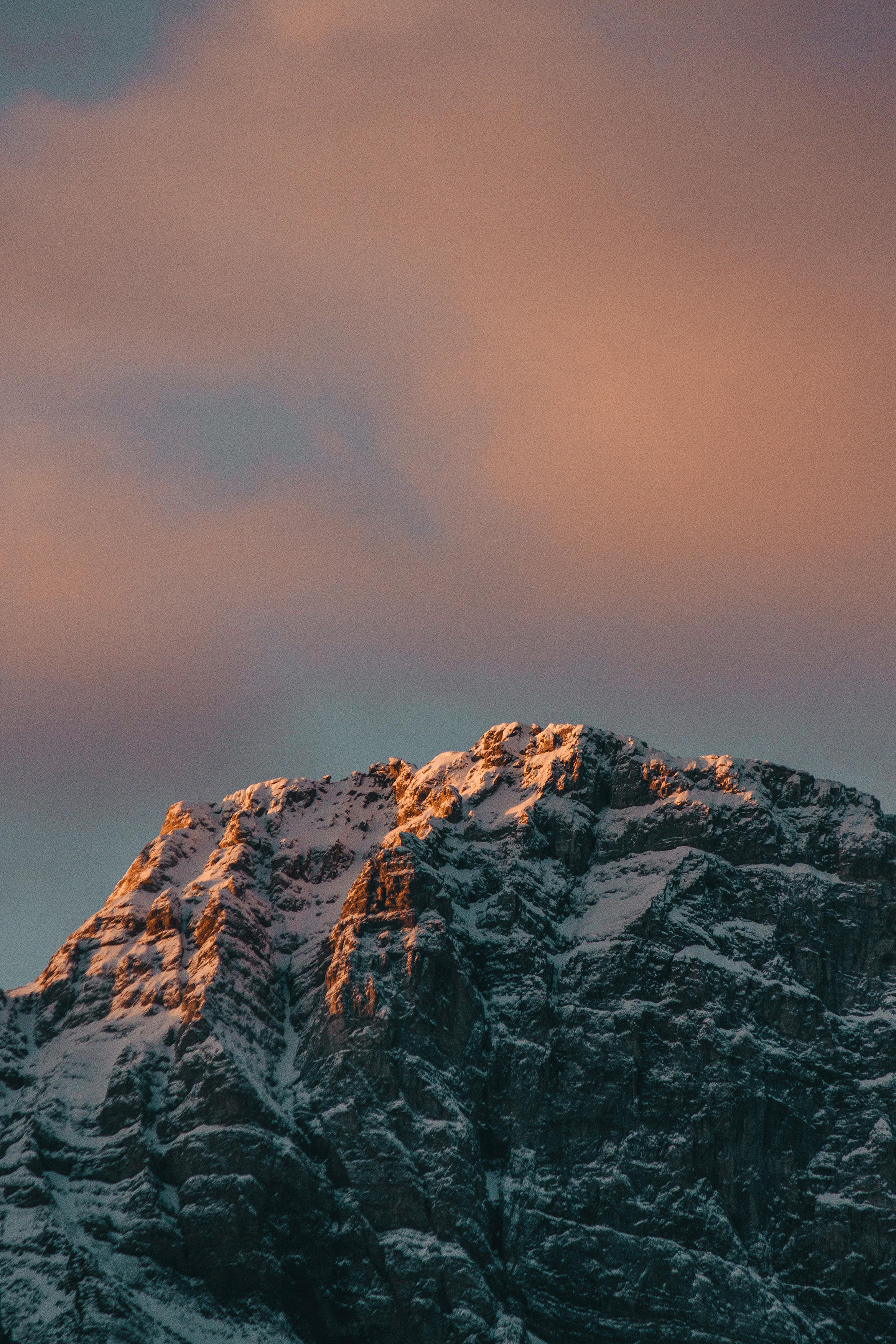 Snow-capped mountain peak bathed in warm sunset light under a sky with pink and orange clouds.