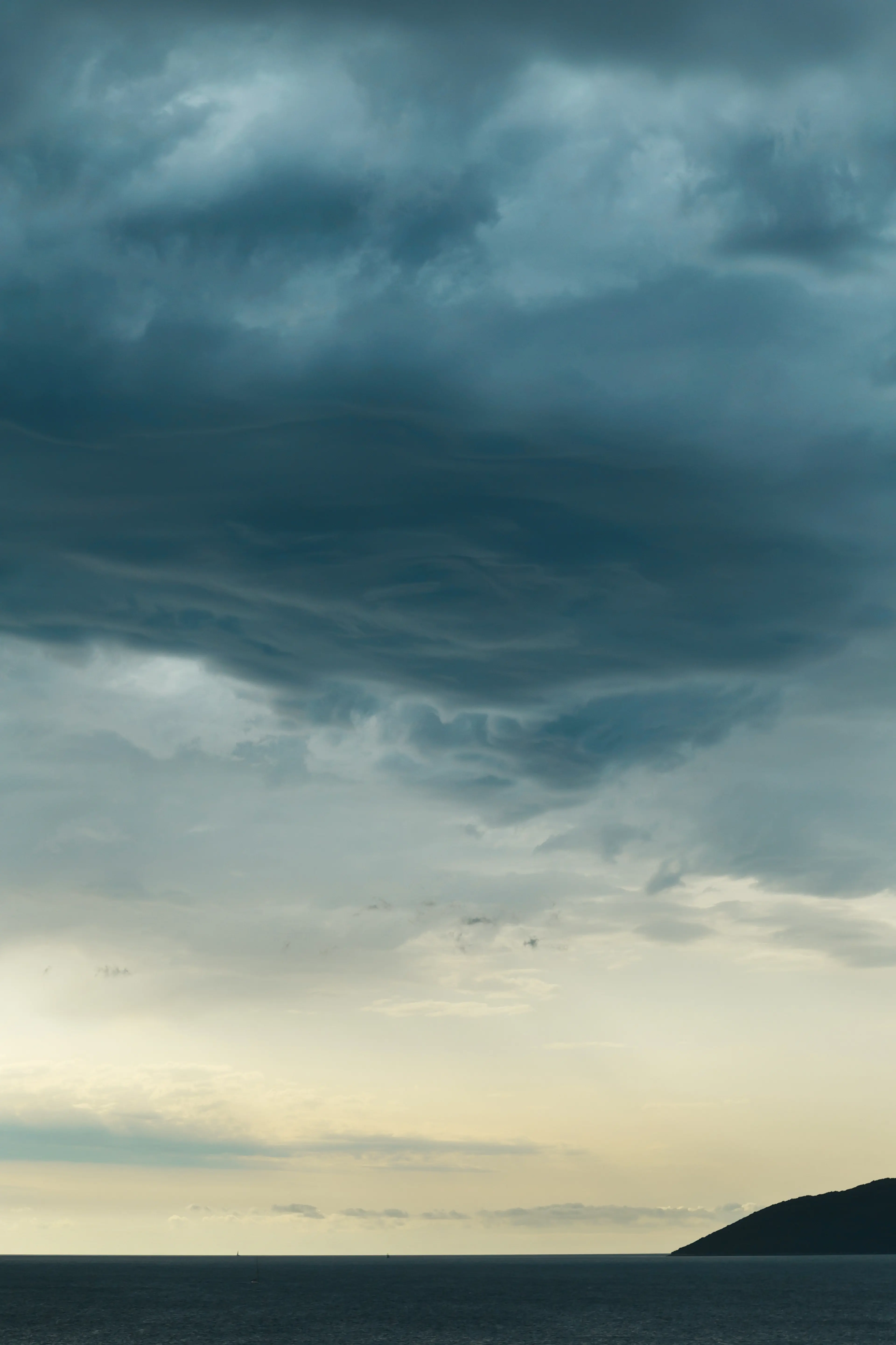 Dramatic cloudy sky over a calm sea horizon with a silhouette of a hill on the right, during a dim sunset.