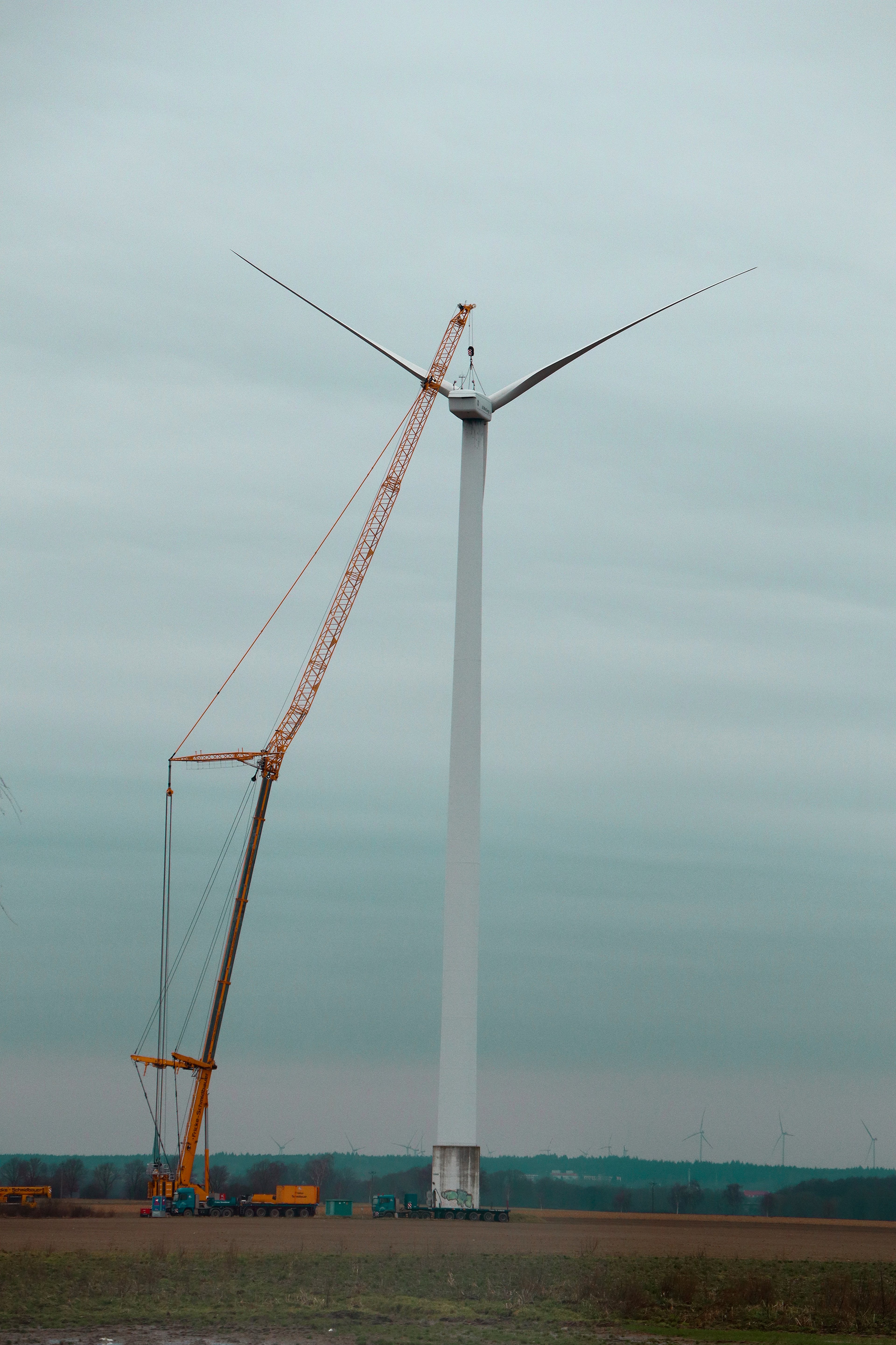 A large crane installing a blade on a wind turbine in an open field under a cloudy sky.
