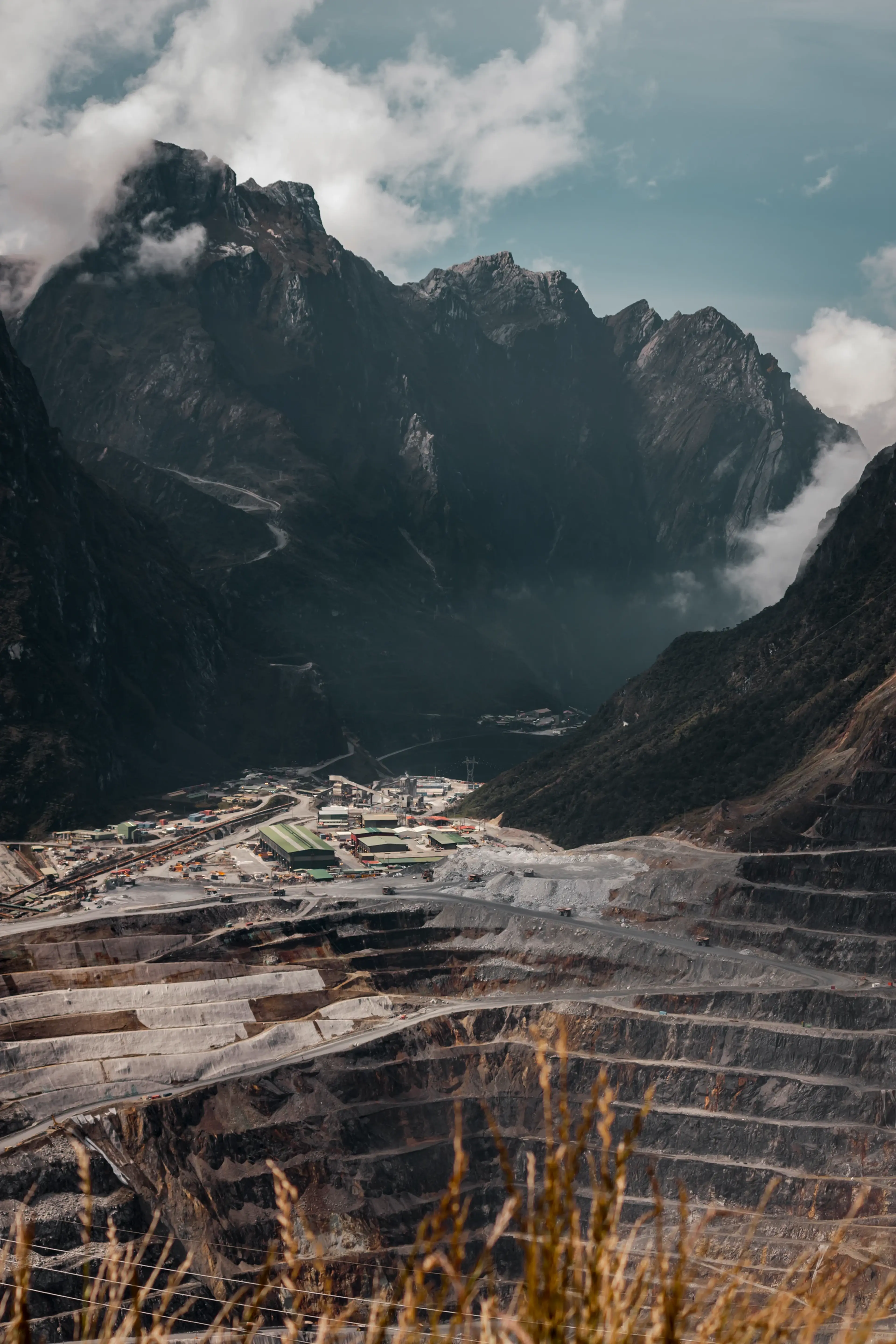 Vast open-pit mine surrounded by rugged mountains with mist and clouds, featuring a settlement and roads leading to the excavation site.