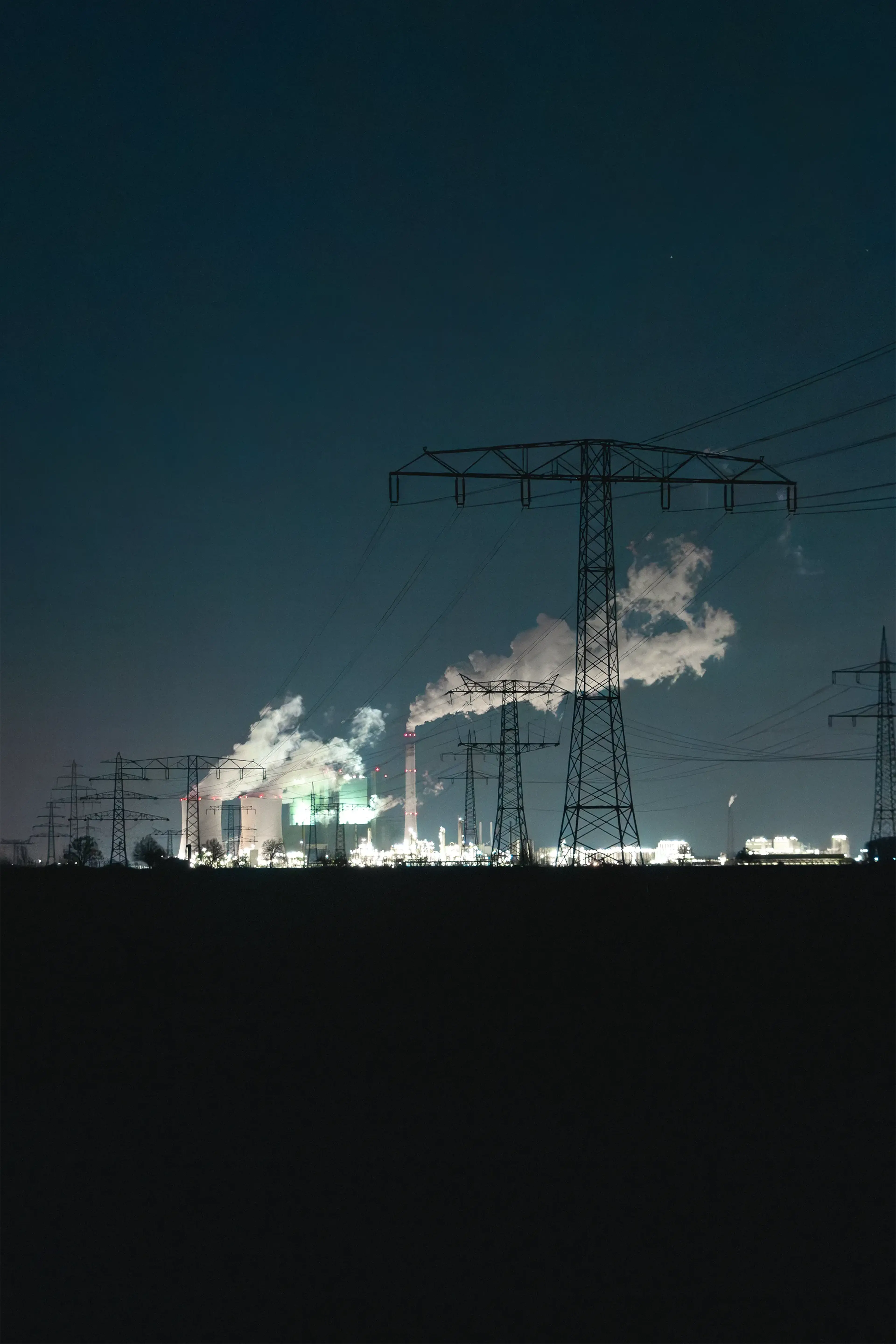 Nighttime industrial scene with illuminated power plant chimneys emitting steam, silhouetted electrical towers, and a dark sky background.