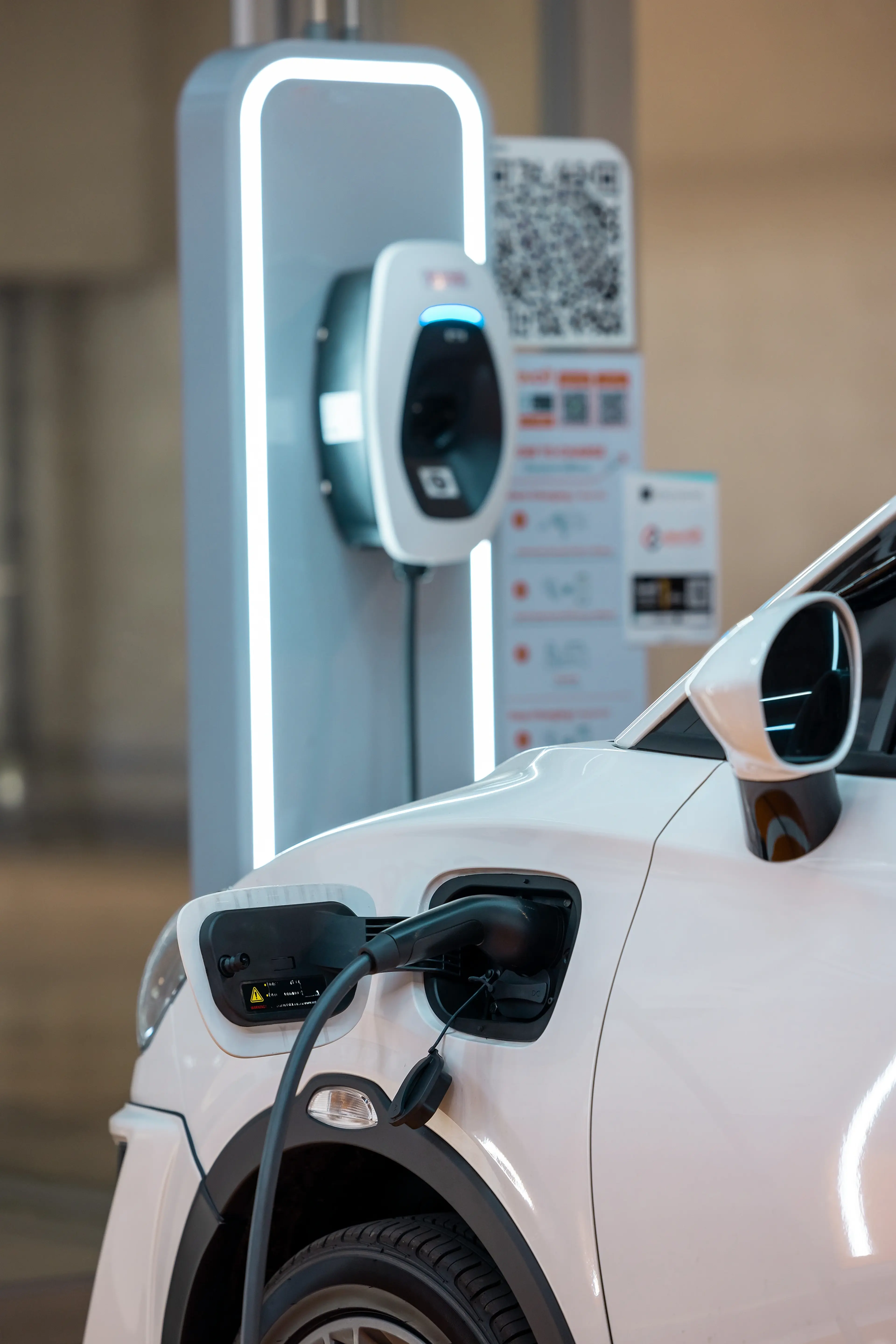 White electric car charging at a station, with a cable plugged into the side, and a digital display on the charger.