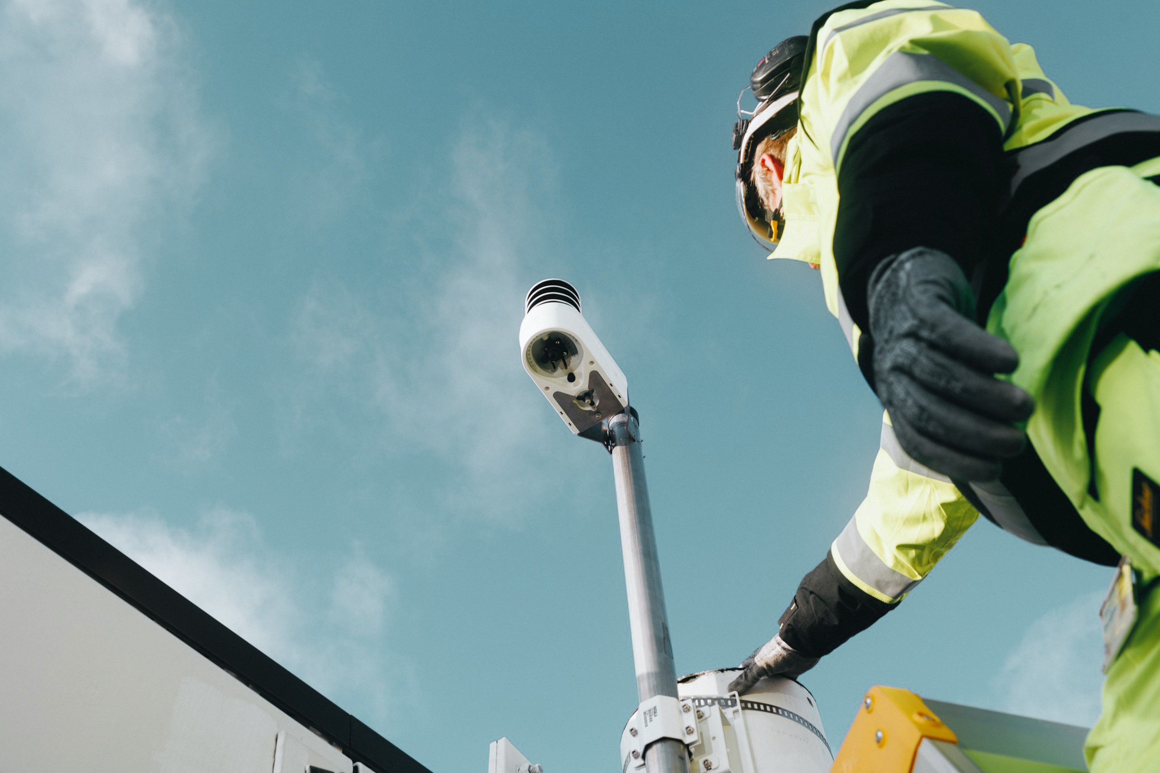 Worker in high-visibility gear adjusting a Vaisala Xweather TempCast weather sensor mounted on a pole against a clear blue sky.