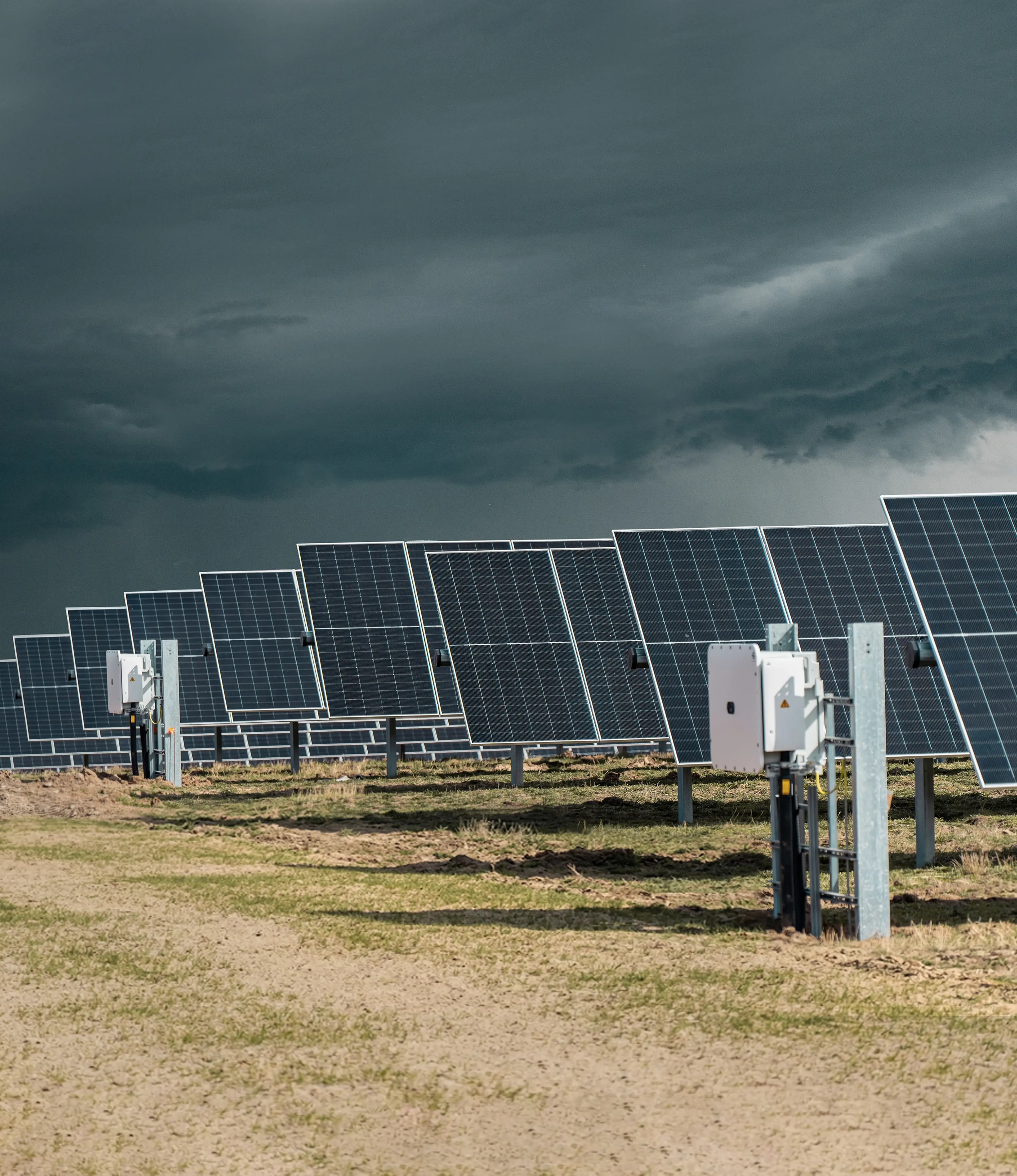 Rows of solar panels under a cloudy sky, installed on a grassy field with electrical equipment visible.