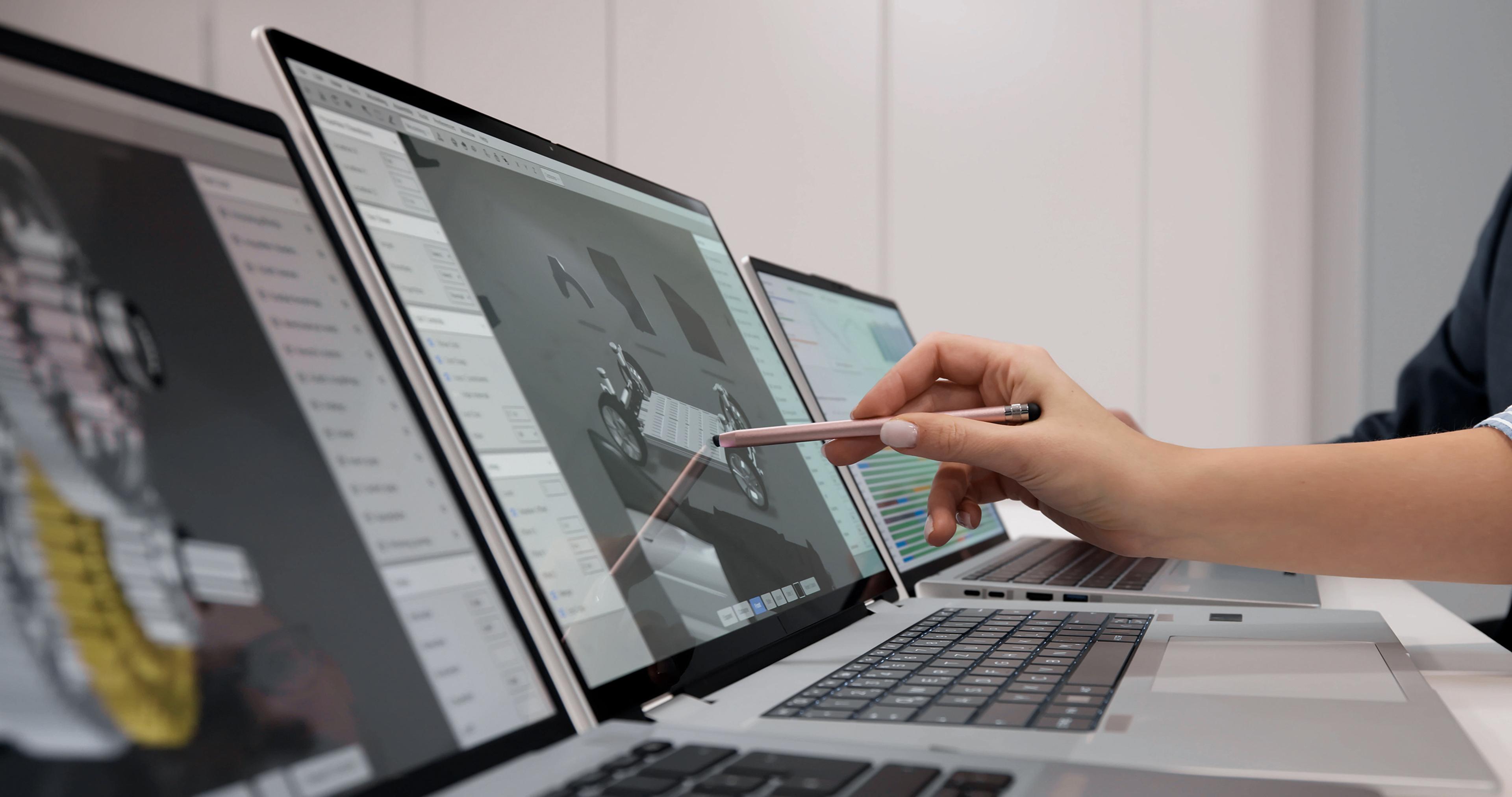 Person using a stylus on a laptop displaying design software, with two other laptops open on a desk in a modern workspace.