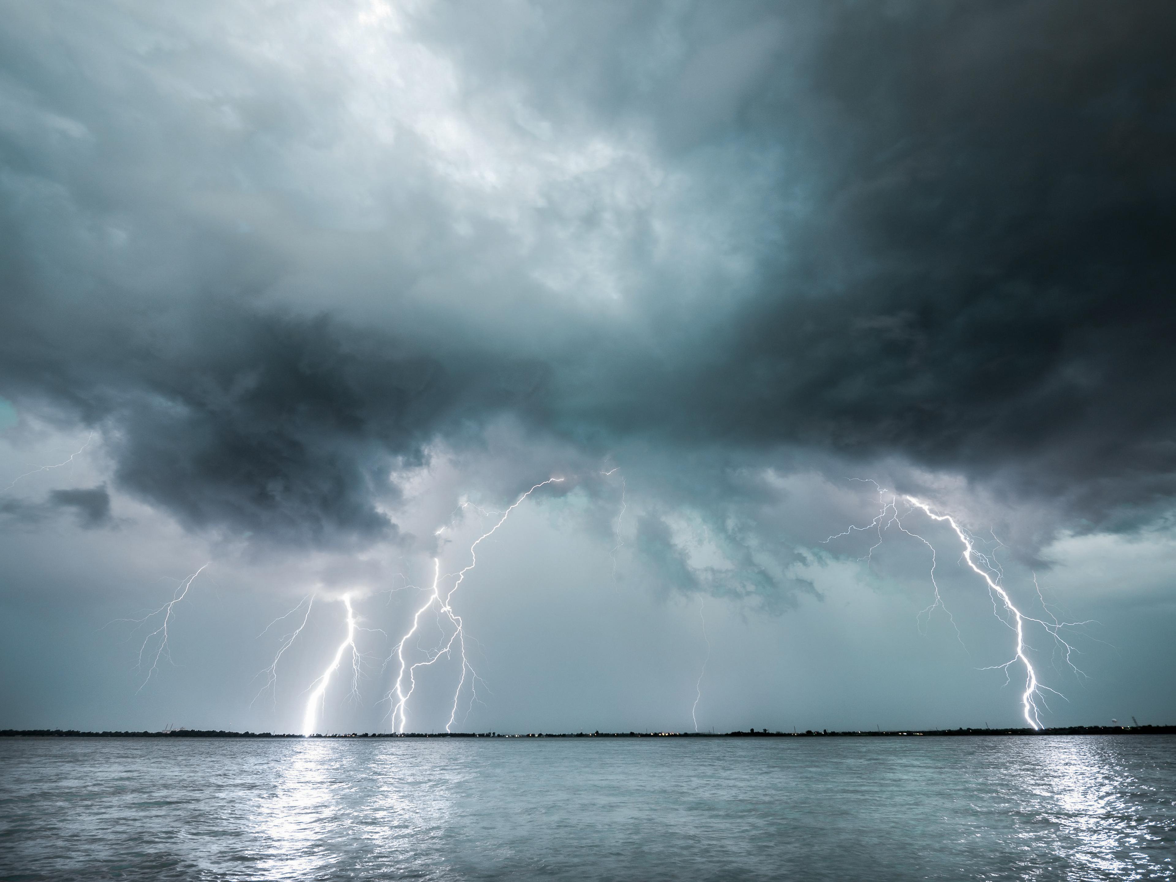 Dramatic lightning storm over a vast body of water, with multiple bright bolts striking from dark, heavy clouds.
