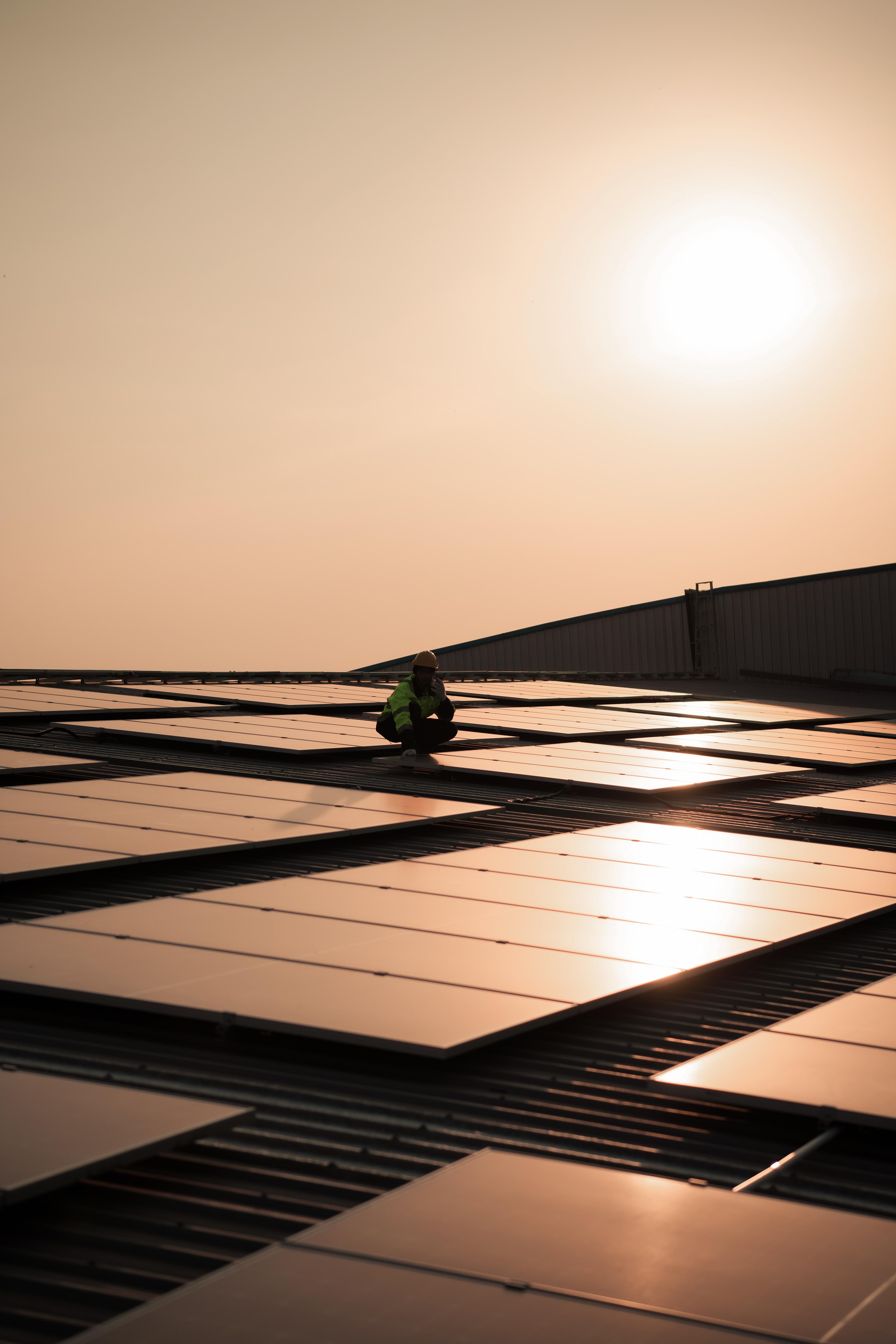 Person working on solar panels on a rooftop at sunset, with a warm orange sky and the sun in the background.