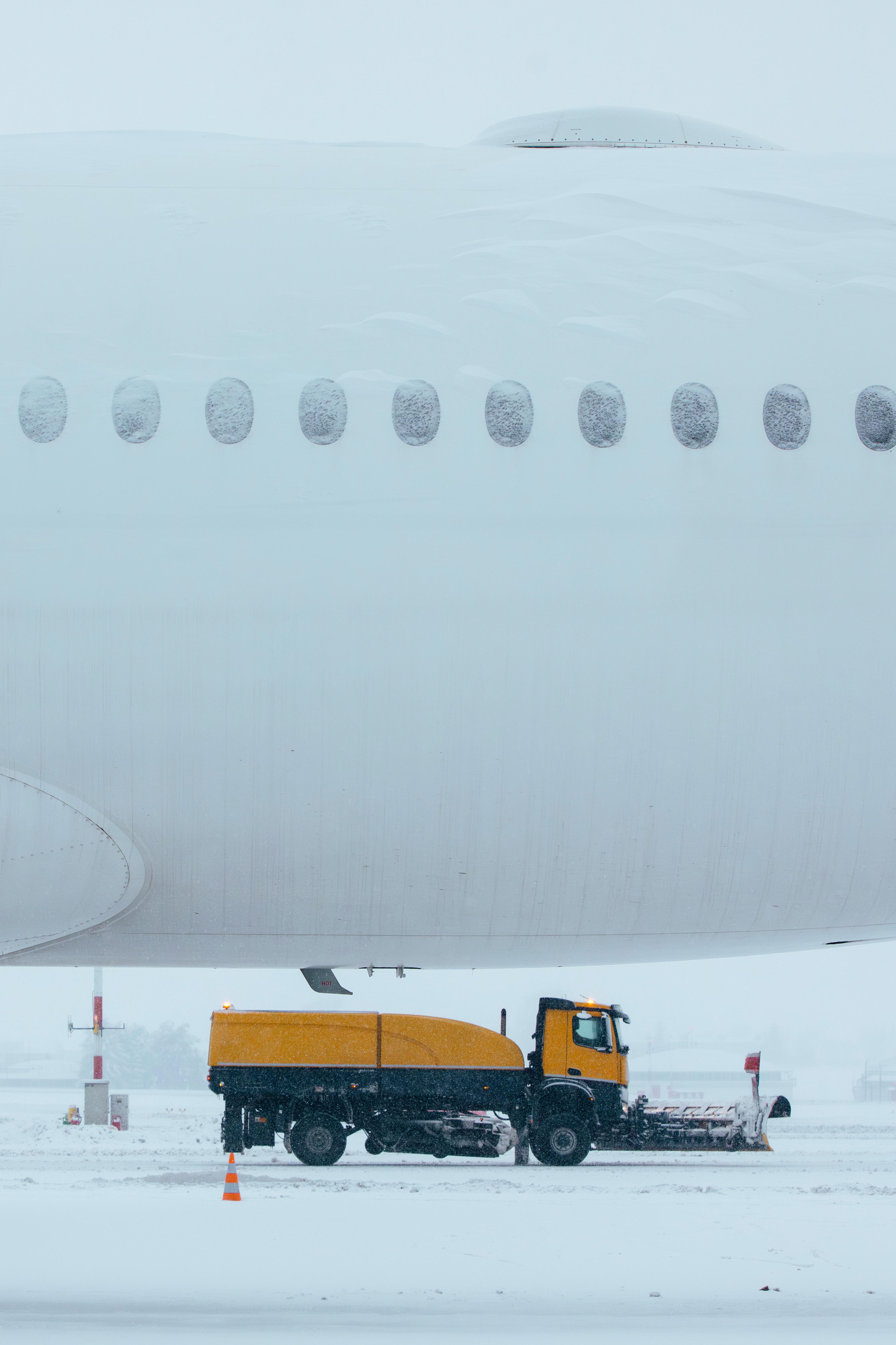 A snowplow truck clears snow beneath the wing of a large airplane on a snowy airport runway.