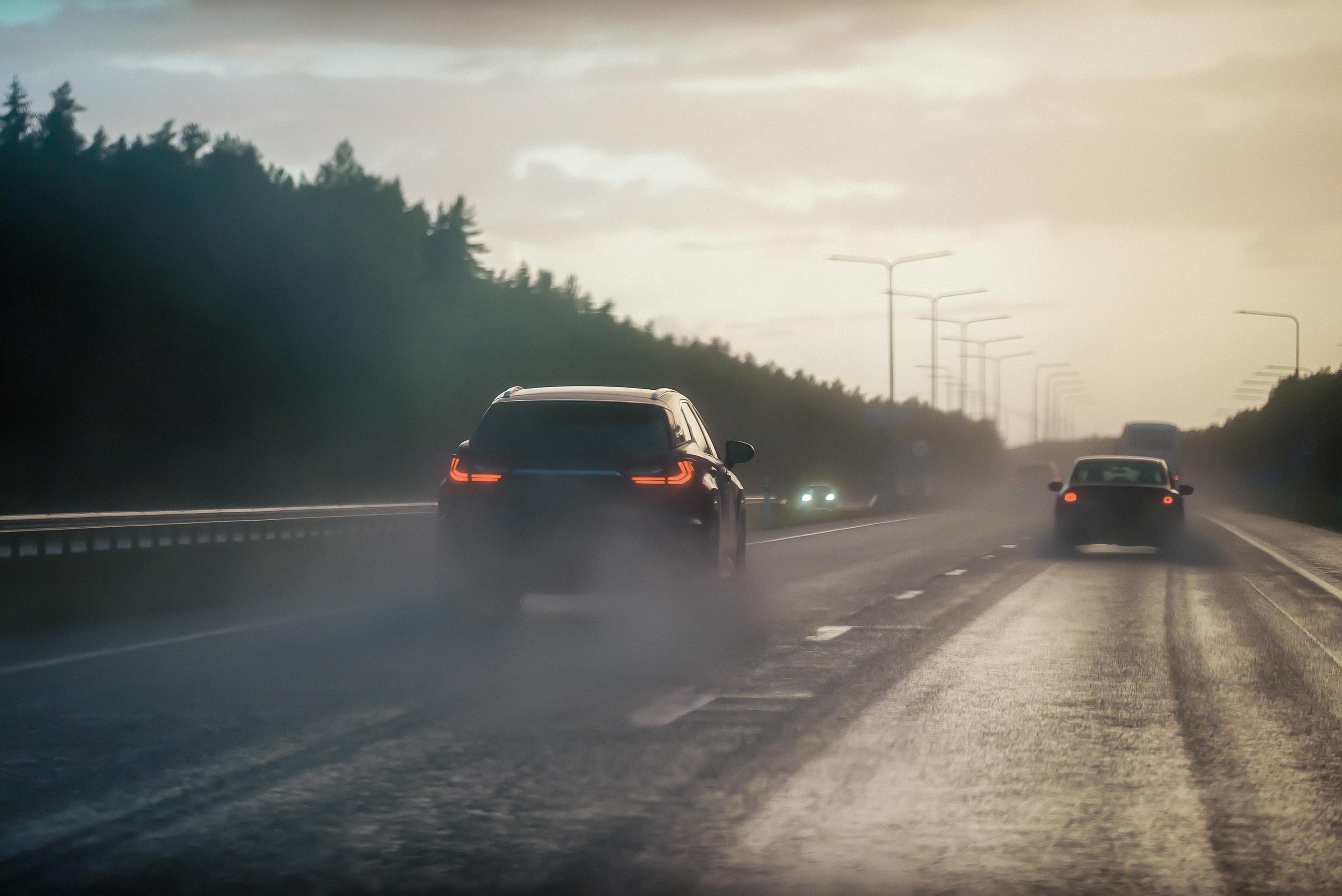 Two cars driving on a wet highway at dusk, with headlights on, surrounded by trees and a cloudy sky.