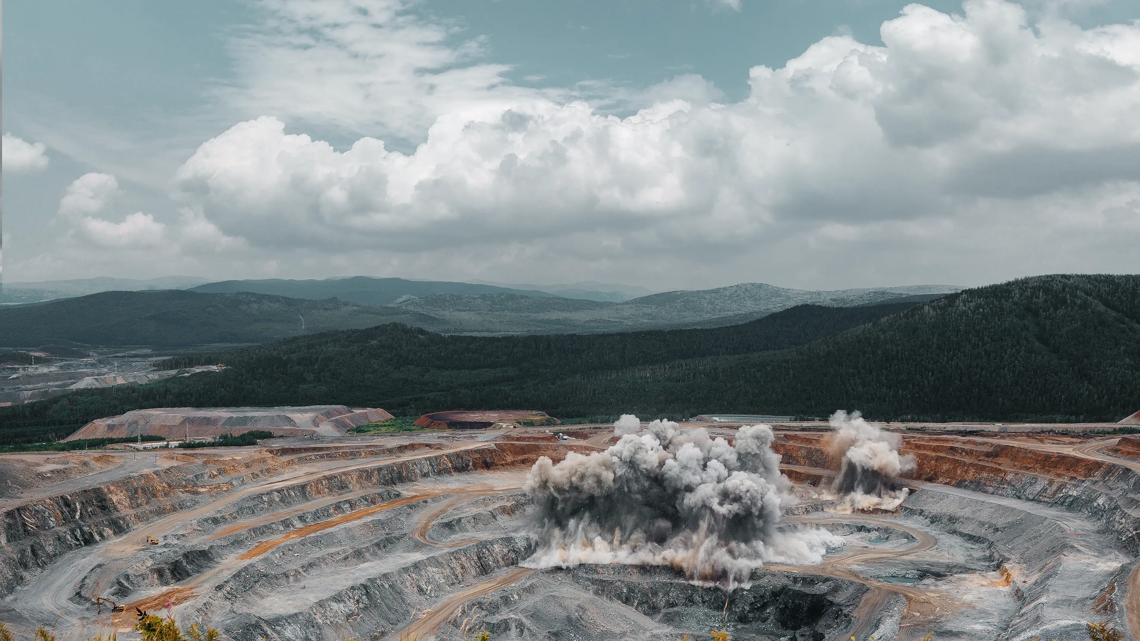 A large open-pit mine with a blast detonation, smoke plumes rising into the air, surrounded by forested hills and a cloudy sky.