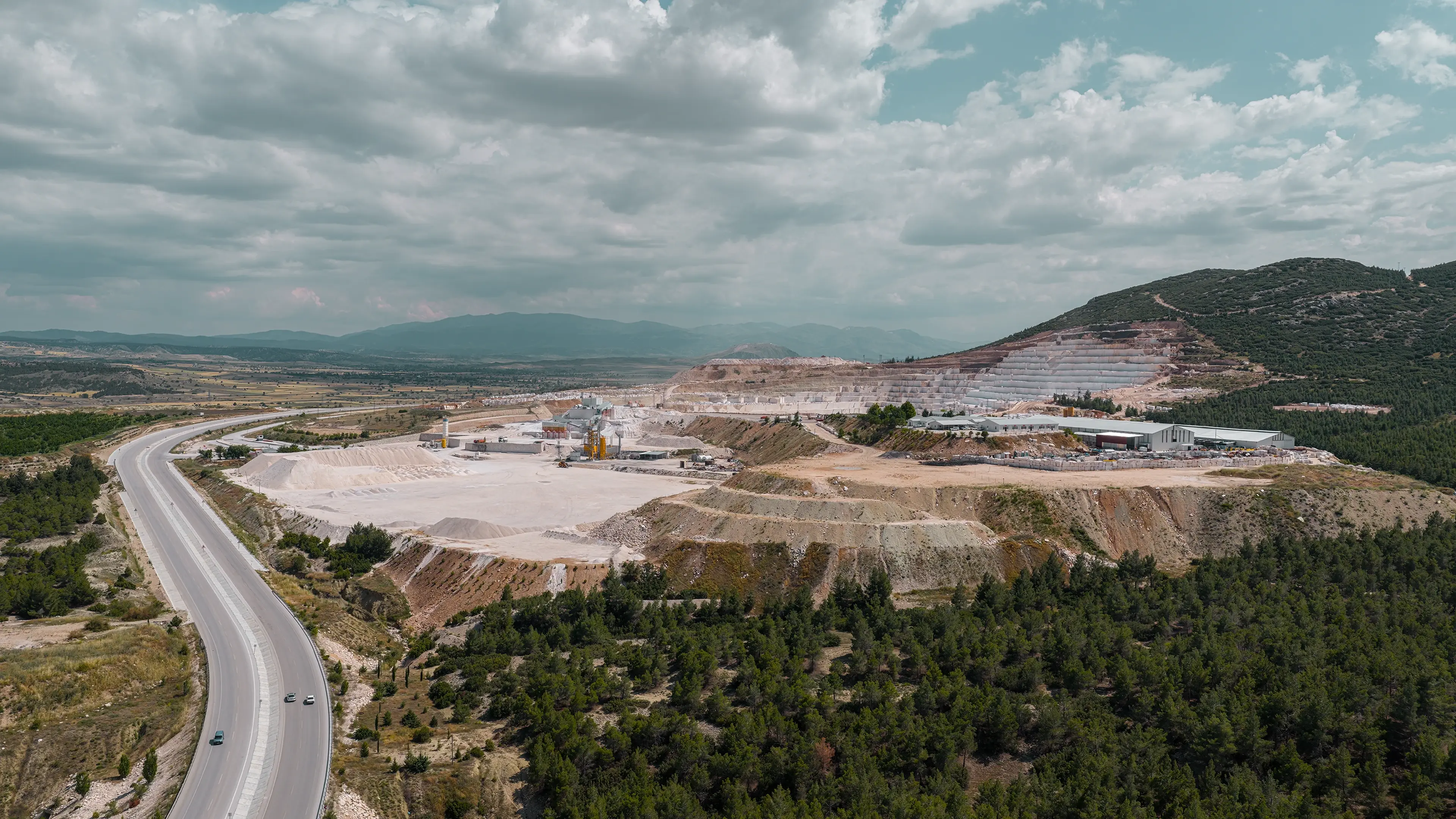 Aerial view of a quarry surrounded by forested hills and a winding road, under a cloudy sky. The landscape extends into the distance.