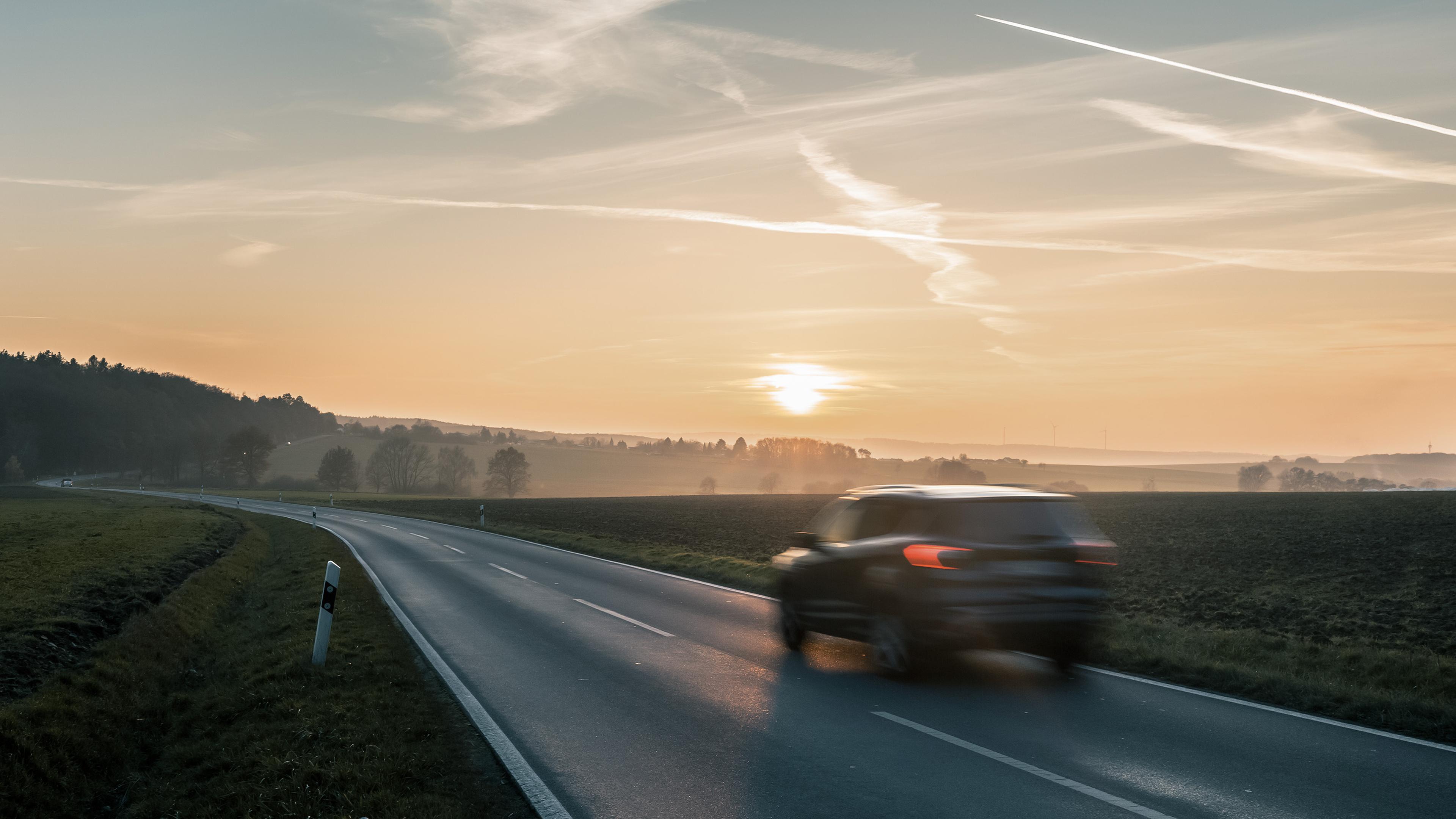 A blurred car drives on a rural road at sunrise, with fields on either side and contrails in the sky.